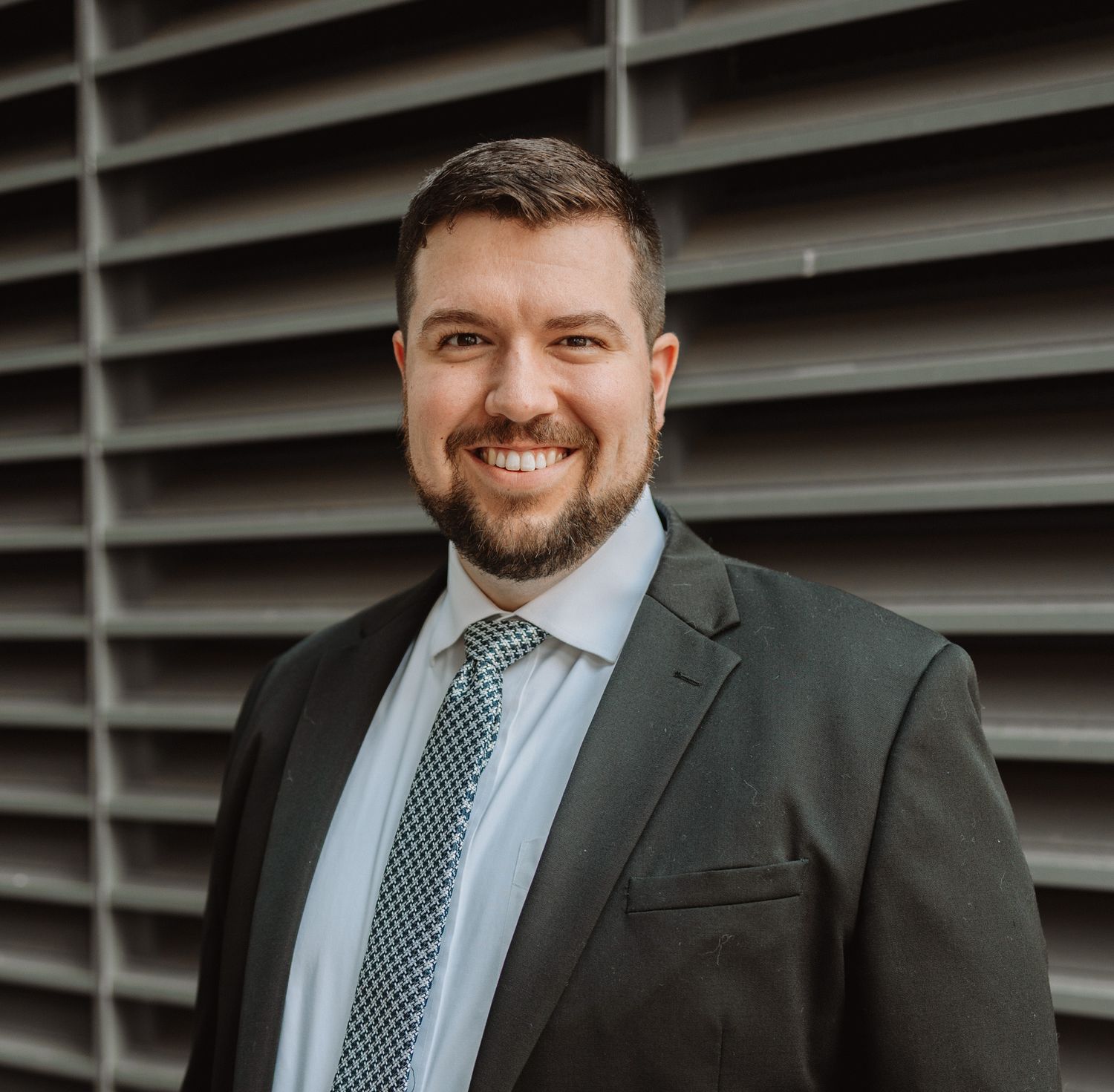 Professional in a dark suit and patterned tie stands against modern architectural background with sleek lines.