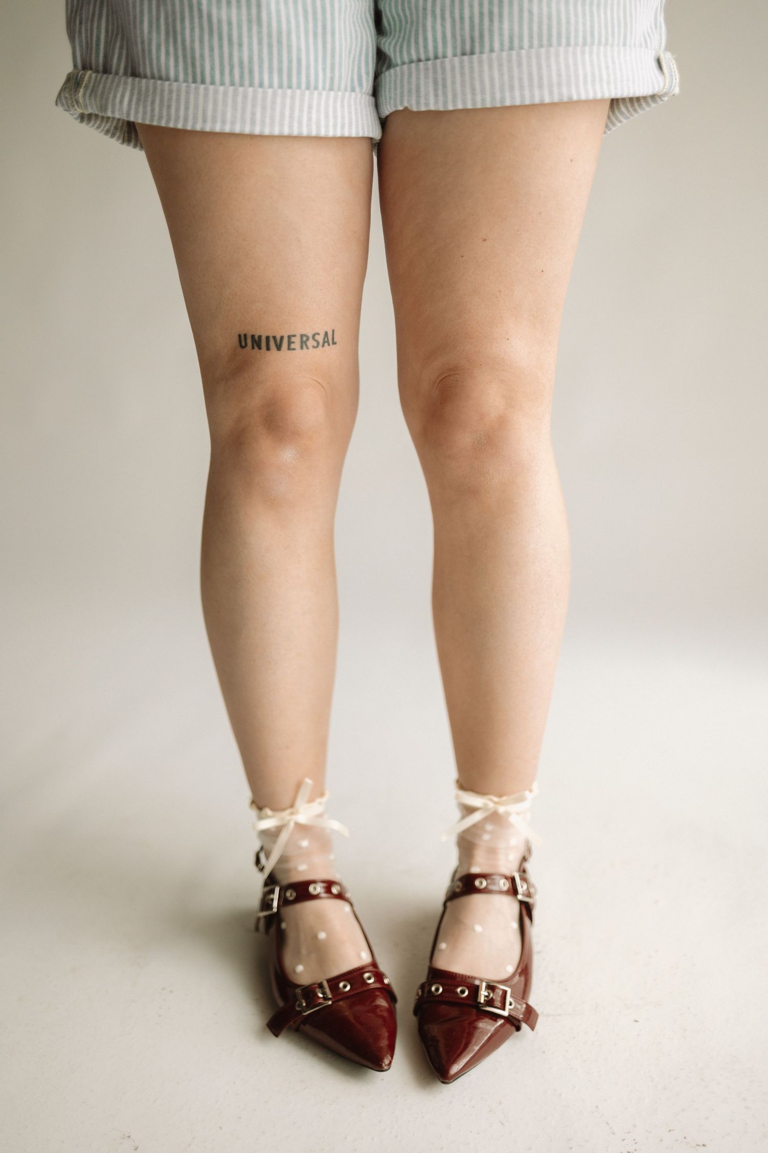 Close-up of legs wearing striped denim shorts, showing 'Universal' tattoo and burgundy strapped ballet flats.