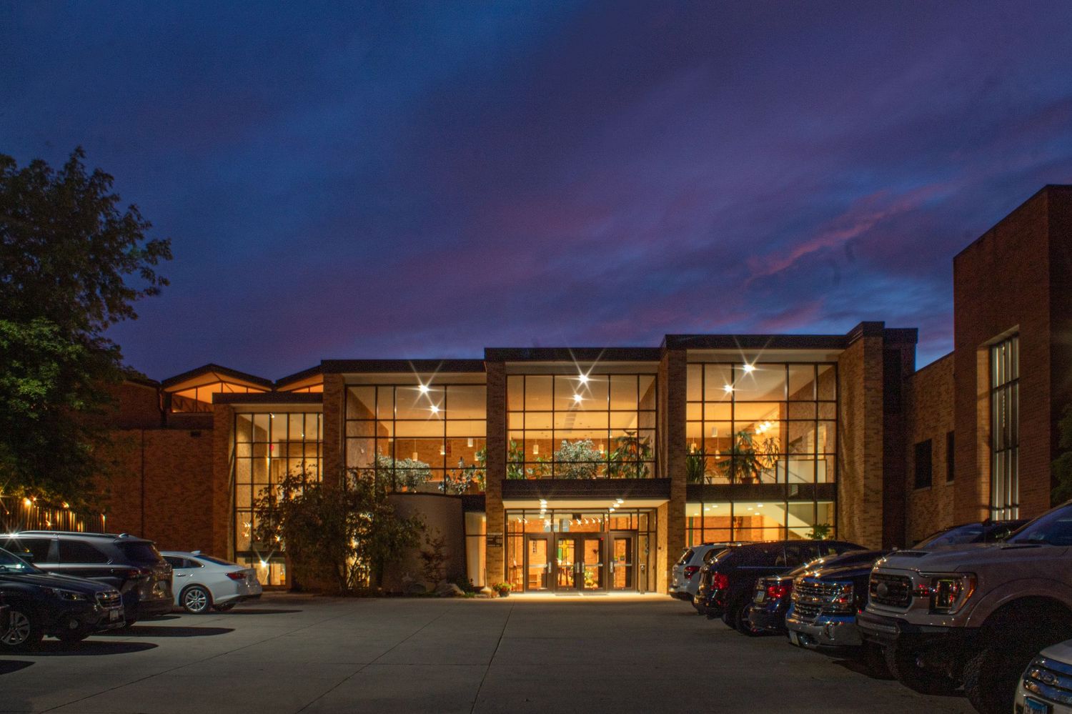 Modern glass-front building illuminated at dusk with warm interior lighting and parked cars in foreground.