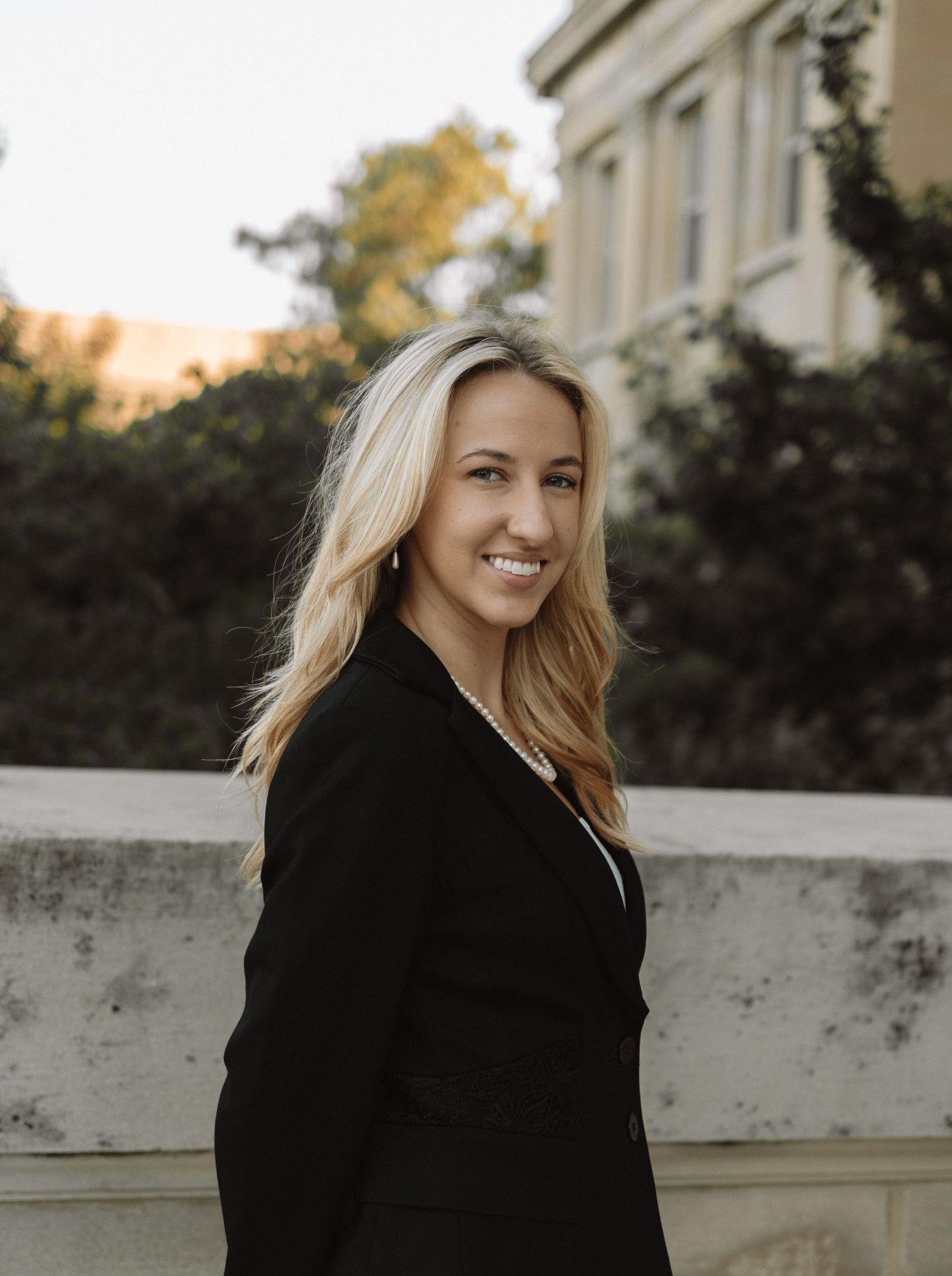 Professional outdoor portrait showing a smiling woman with blonde hair wearing a black blazer against an architectural backdrop.