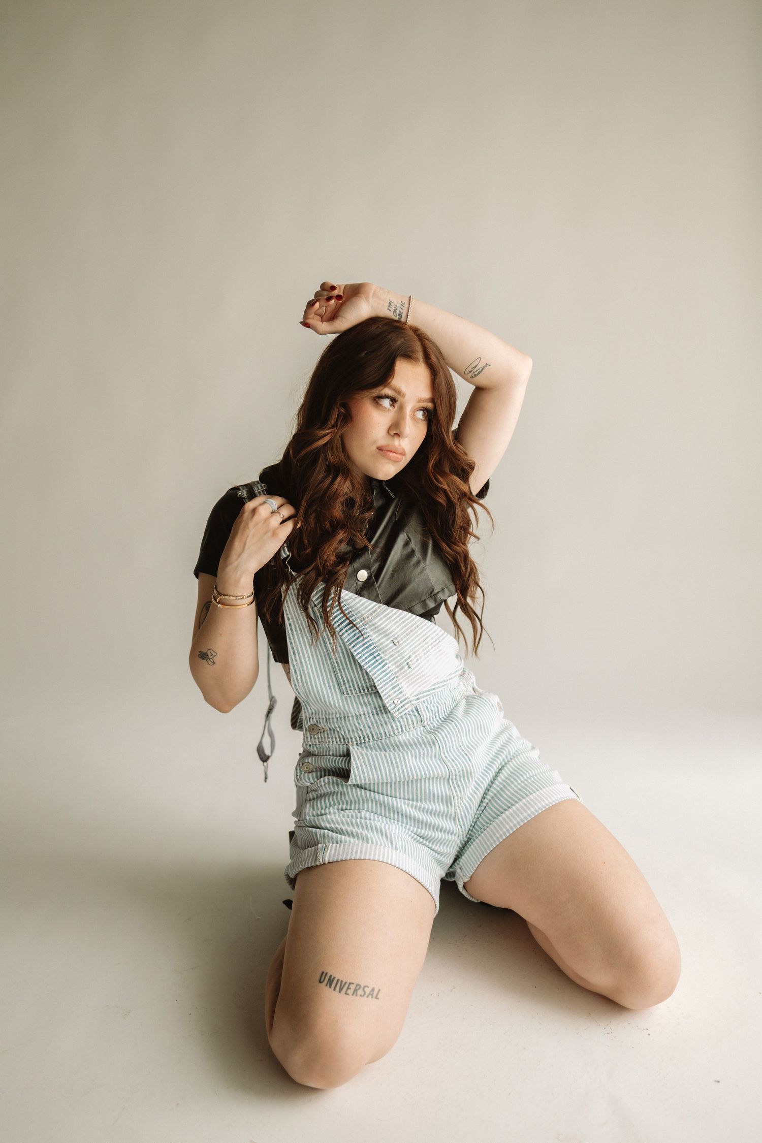Woman in light denim overalls and black top poses on floor against neutral background in artistic portrait session.