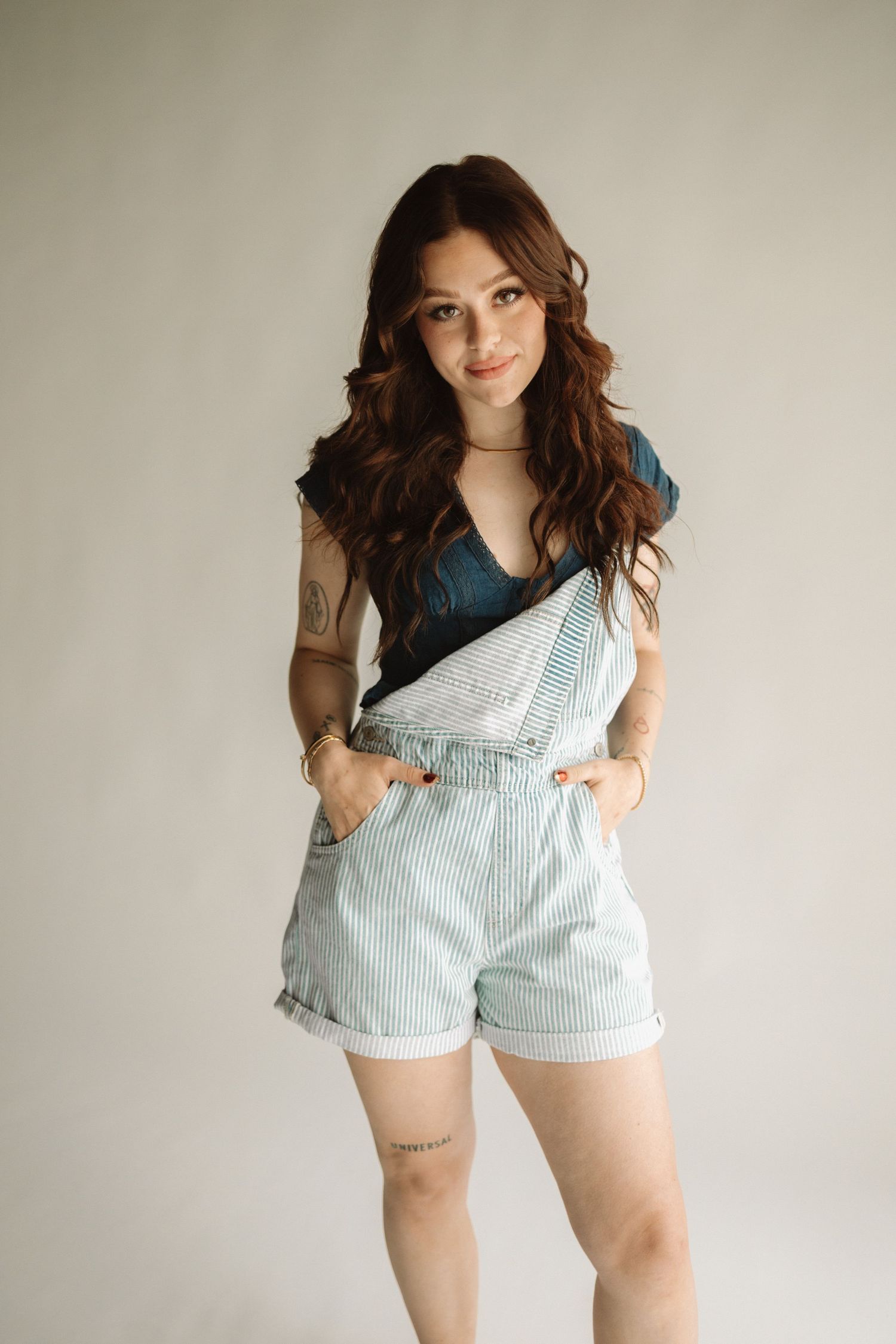 Casual fashion portrait with striped denim overalls shorts and dark top against neutral background.