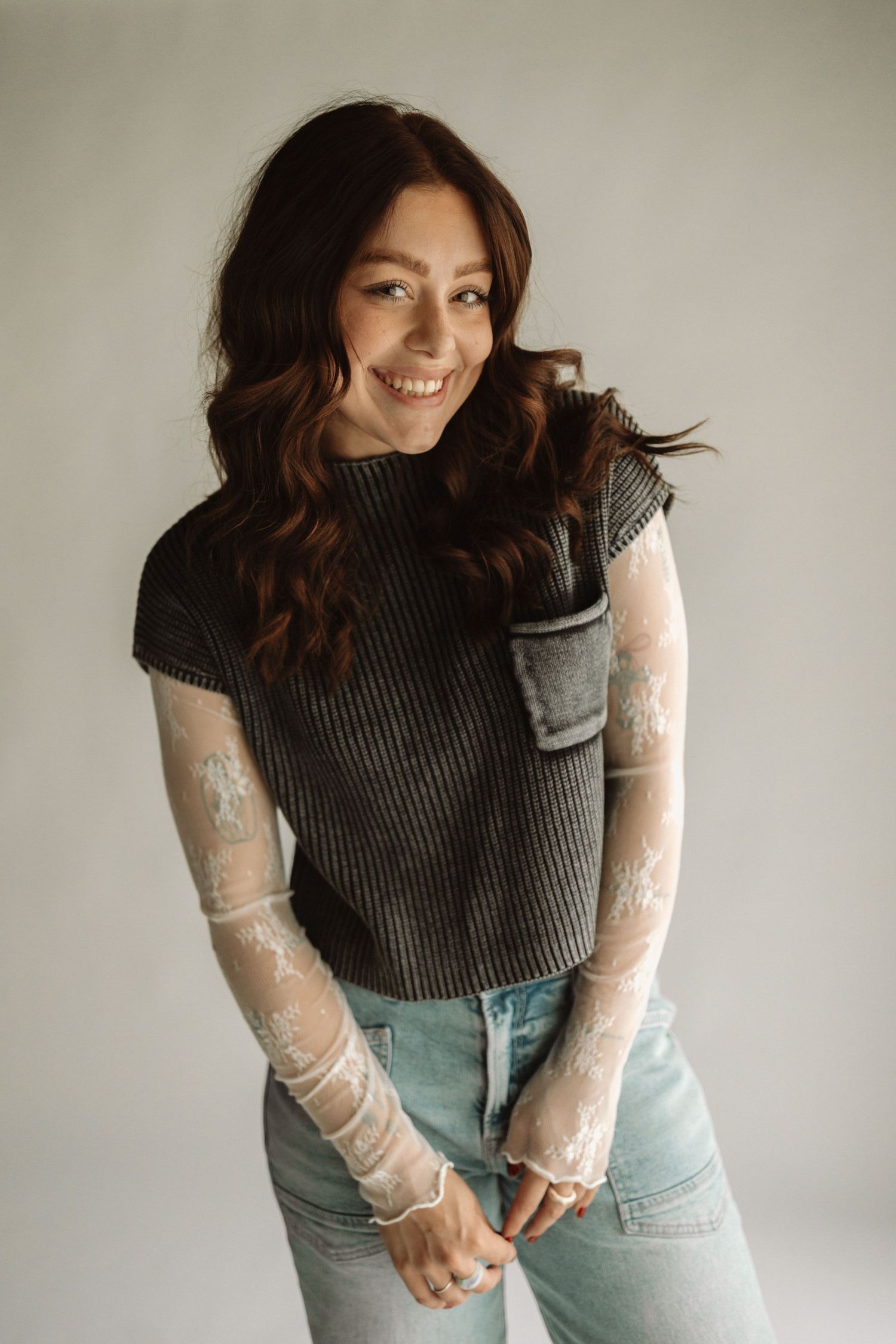 Model in grey sleeveless top with sheer lace sleeves and light denim jeans poses casually against neutral background.