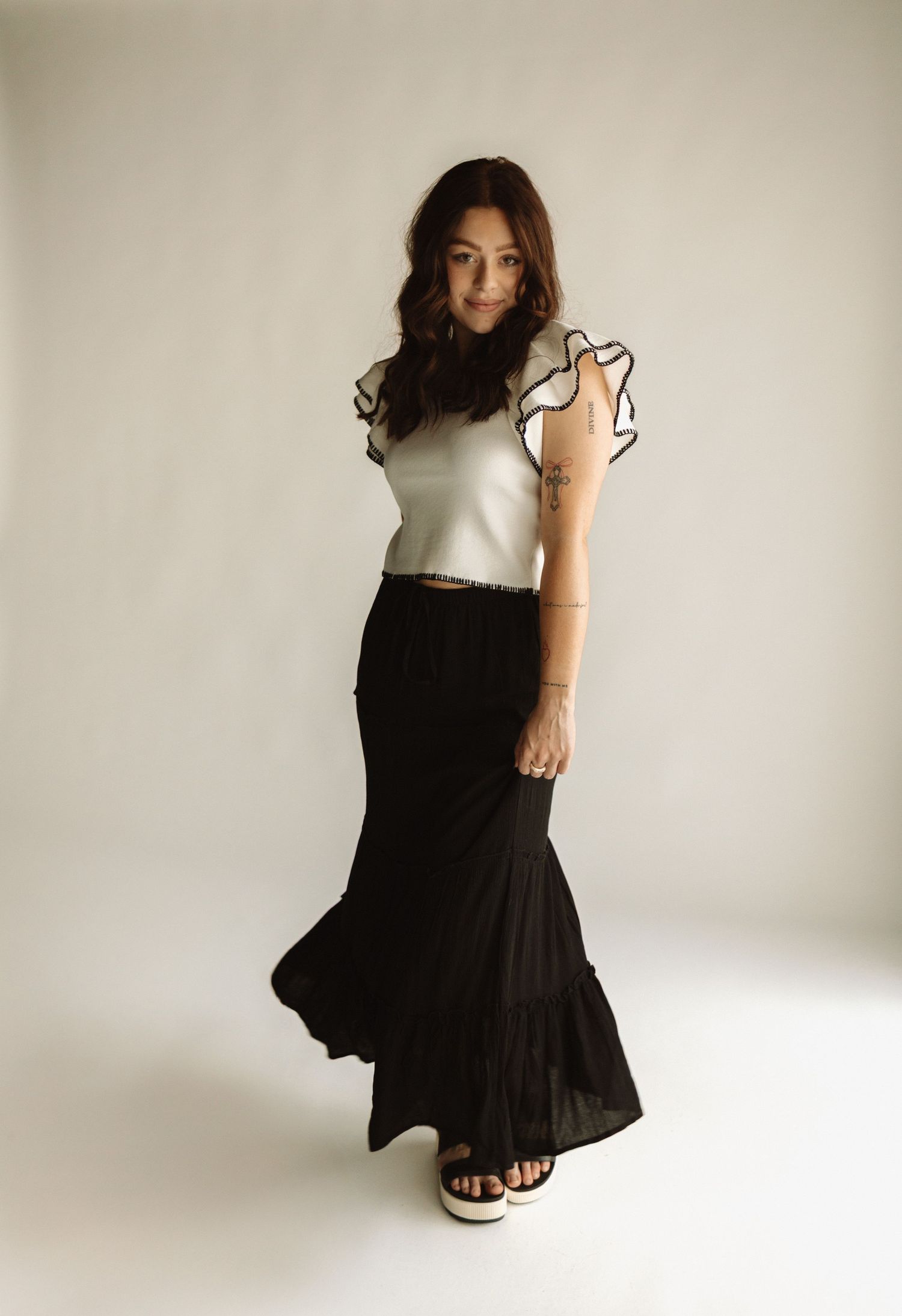 A woman poses in a white ruffled sleeve top and flowing black maxi skirt against a light neutral background.