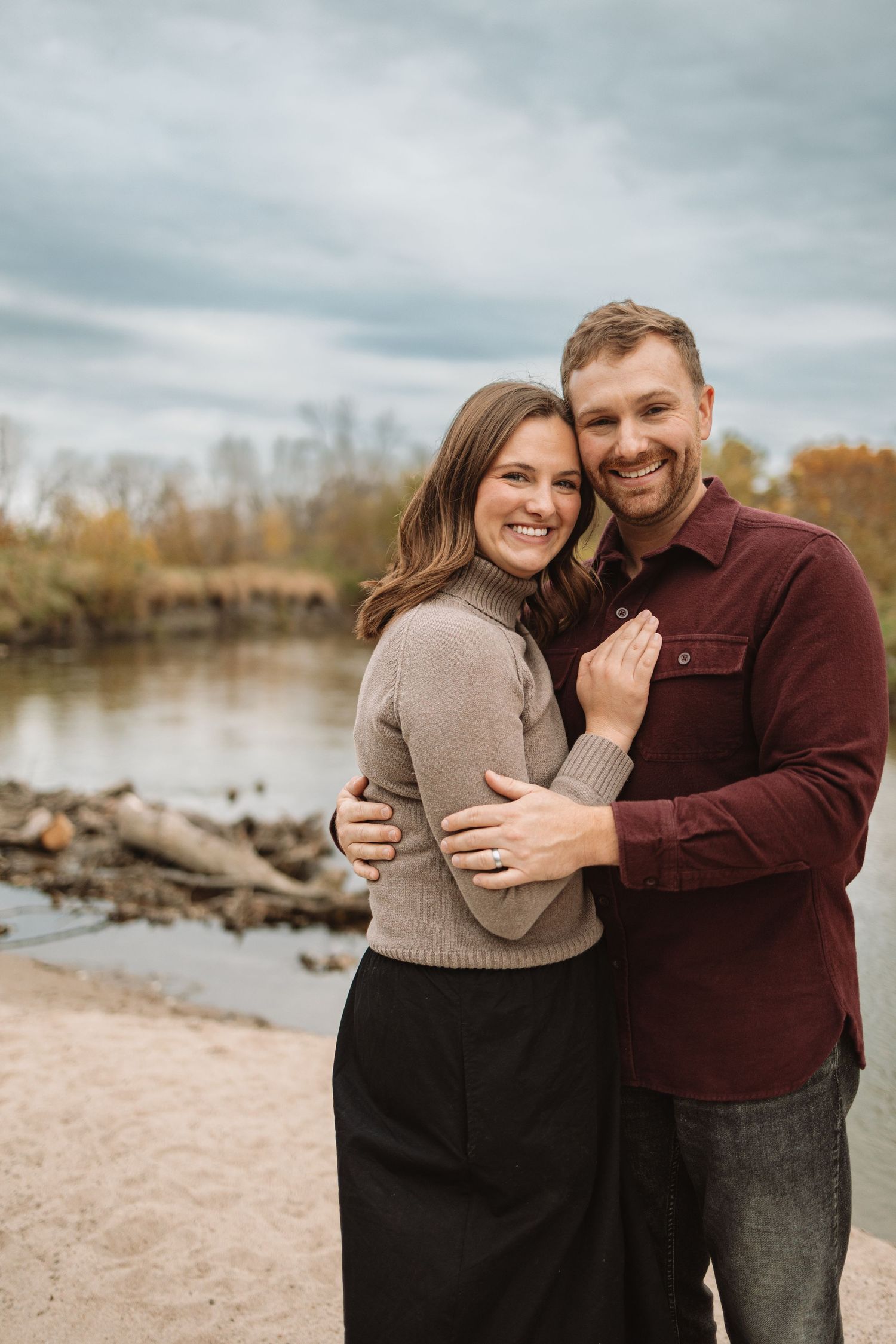 A couple embraces and laughs together during a romantic photoshoot by a lake on an overcast autumn day.