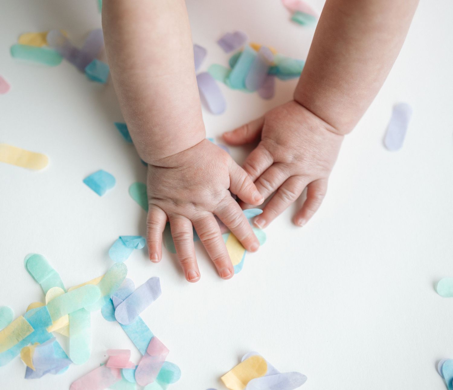 Small baby hands reach down to touch colorful pastel paper confetti scattered on a white surface.
