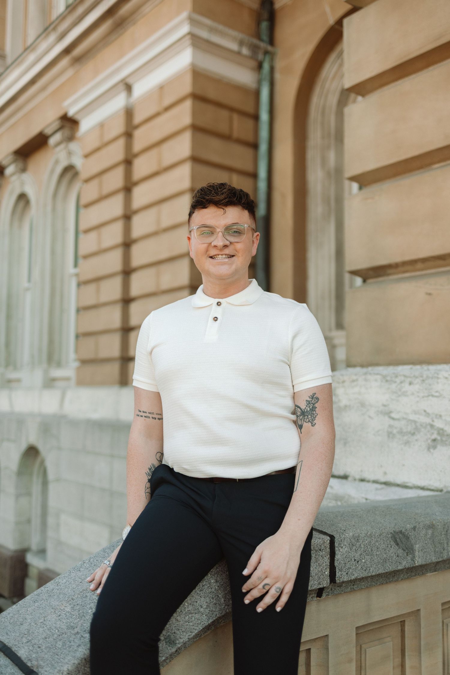 A person in a white polo shirt and dark pants sits casually on a concrete ledge outside a building with stone walls.
