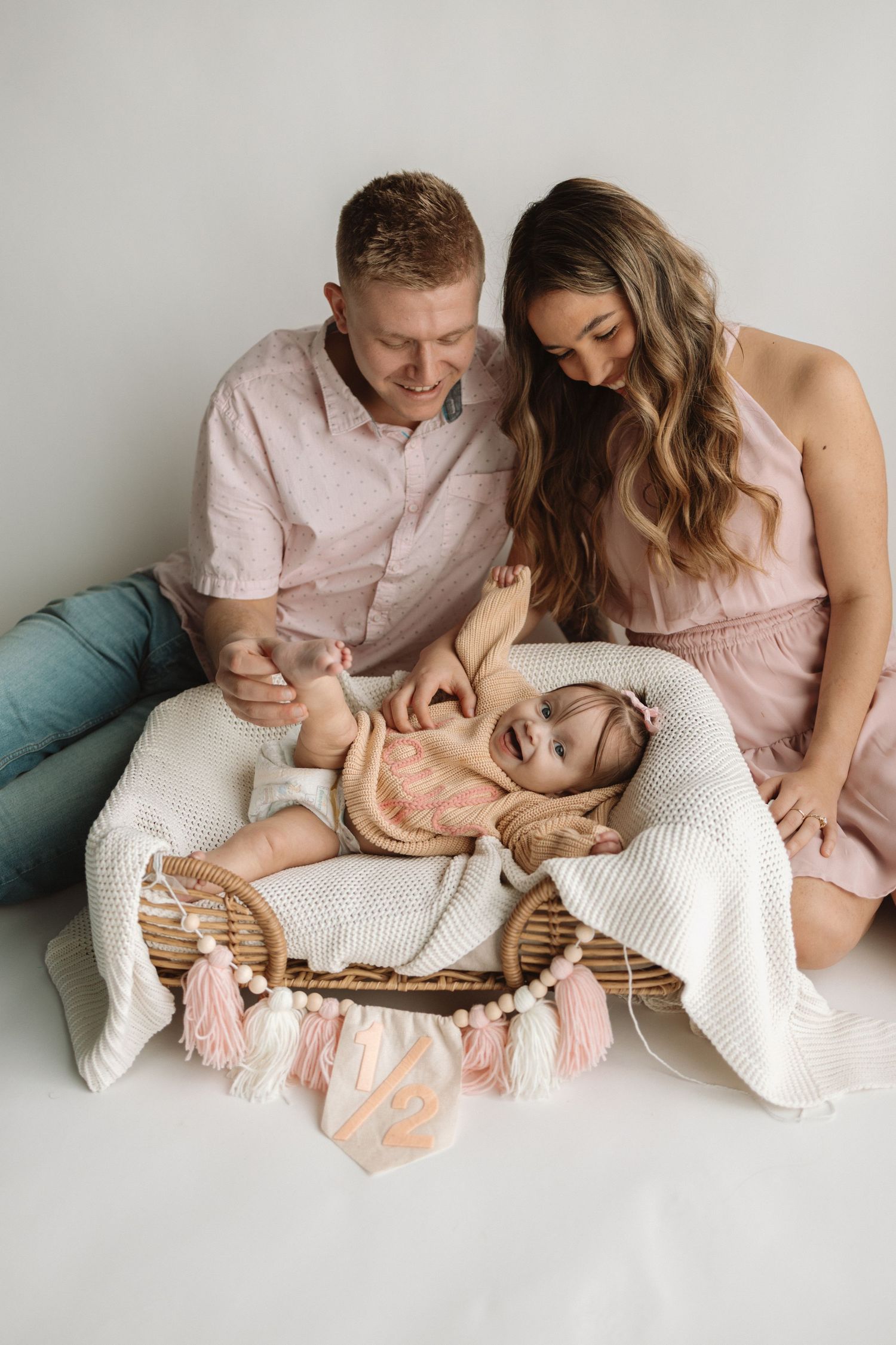 Family in coordinating pink outfits sits together on white backdrop with decorative tasseled banner.