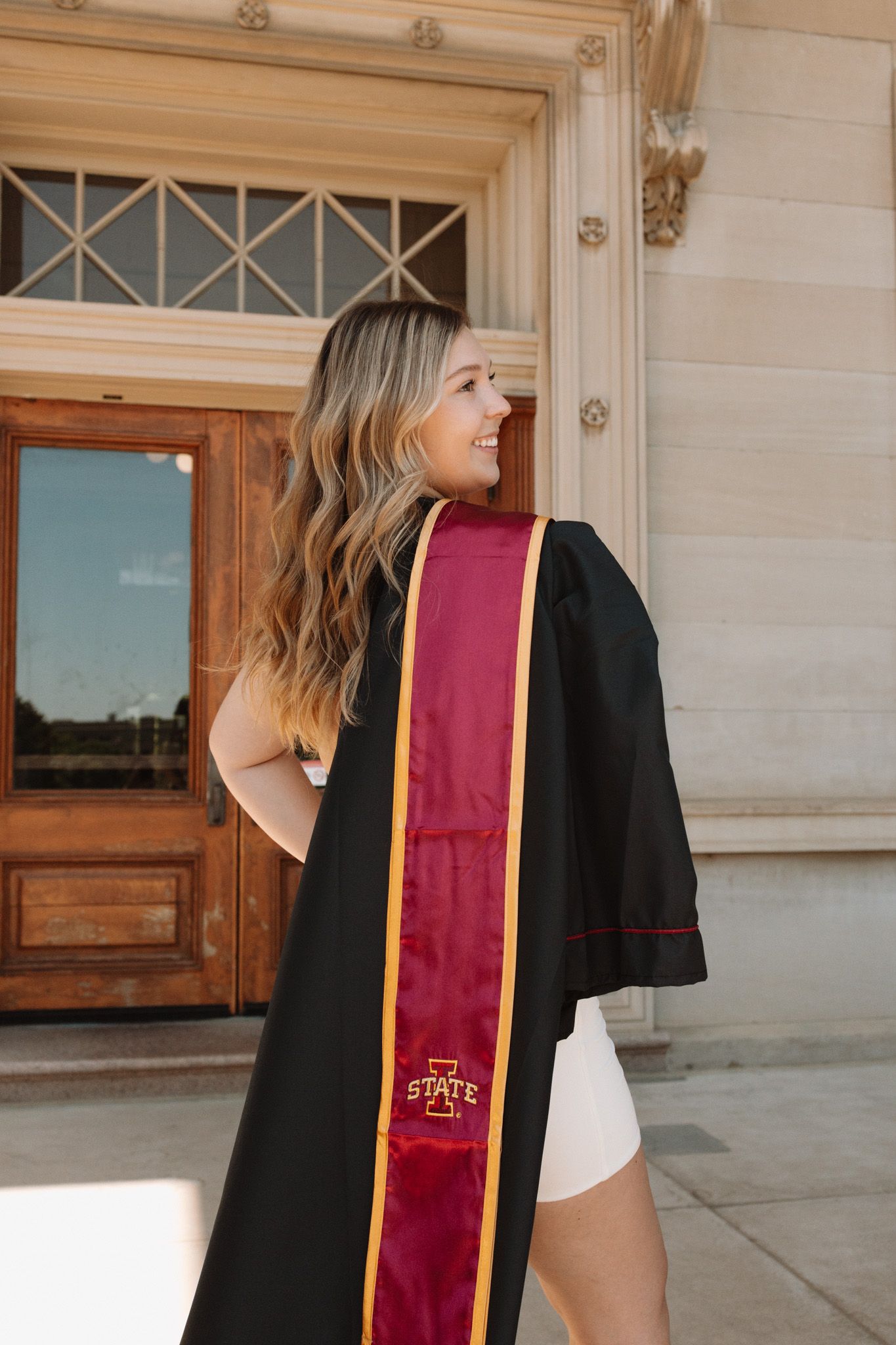 Graduate in black cap and gown with burgundy honor stole walks along stone building in outdoor setting.