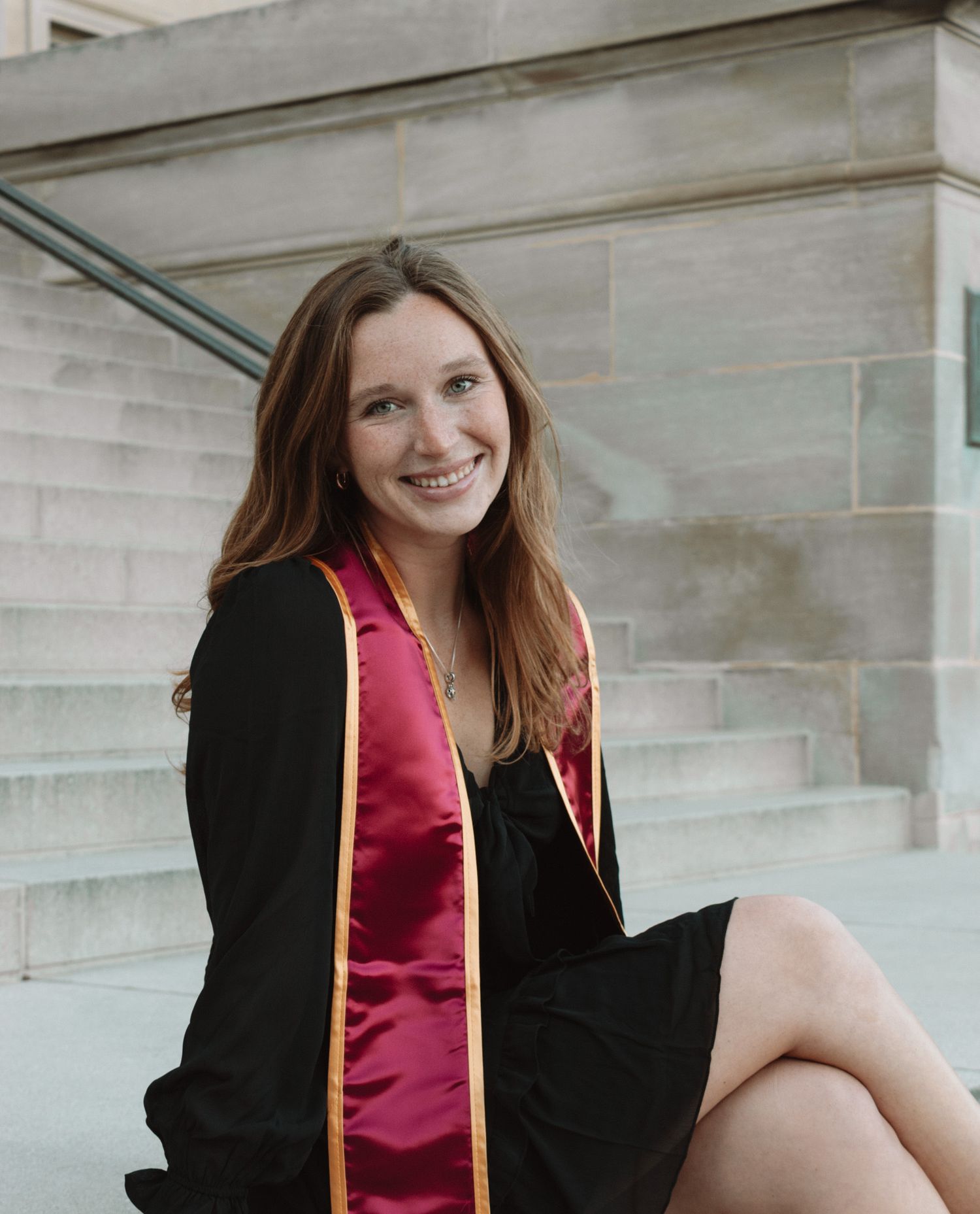 Graduate wearing a red graduation stole and black dress sits on stone steps during a graduation photo session.