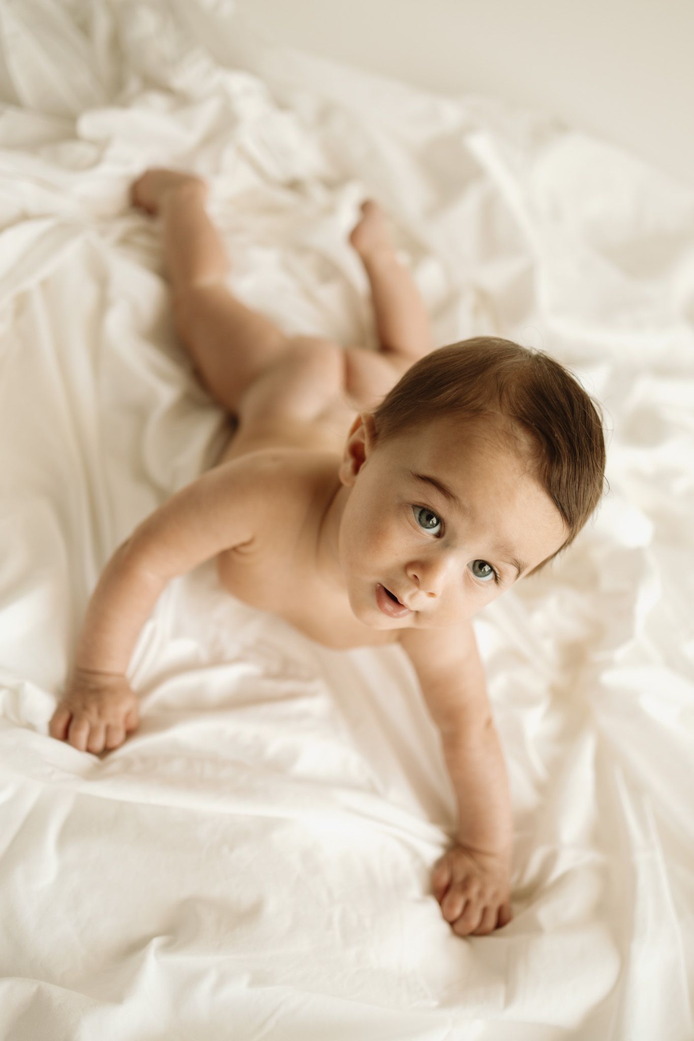 A small baby lying on white bedding looks up at the camera with a gentle expression.