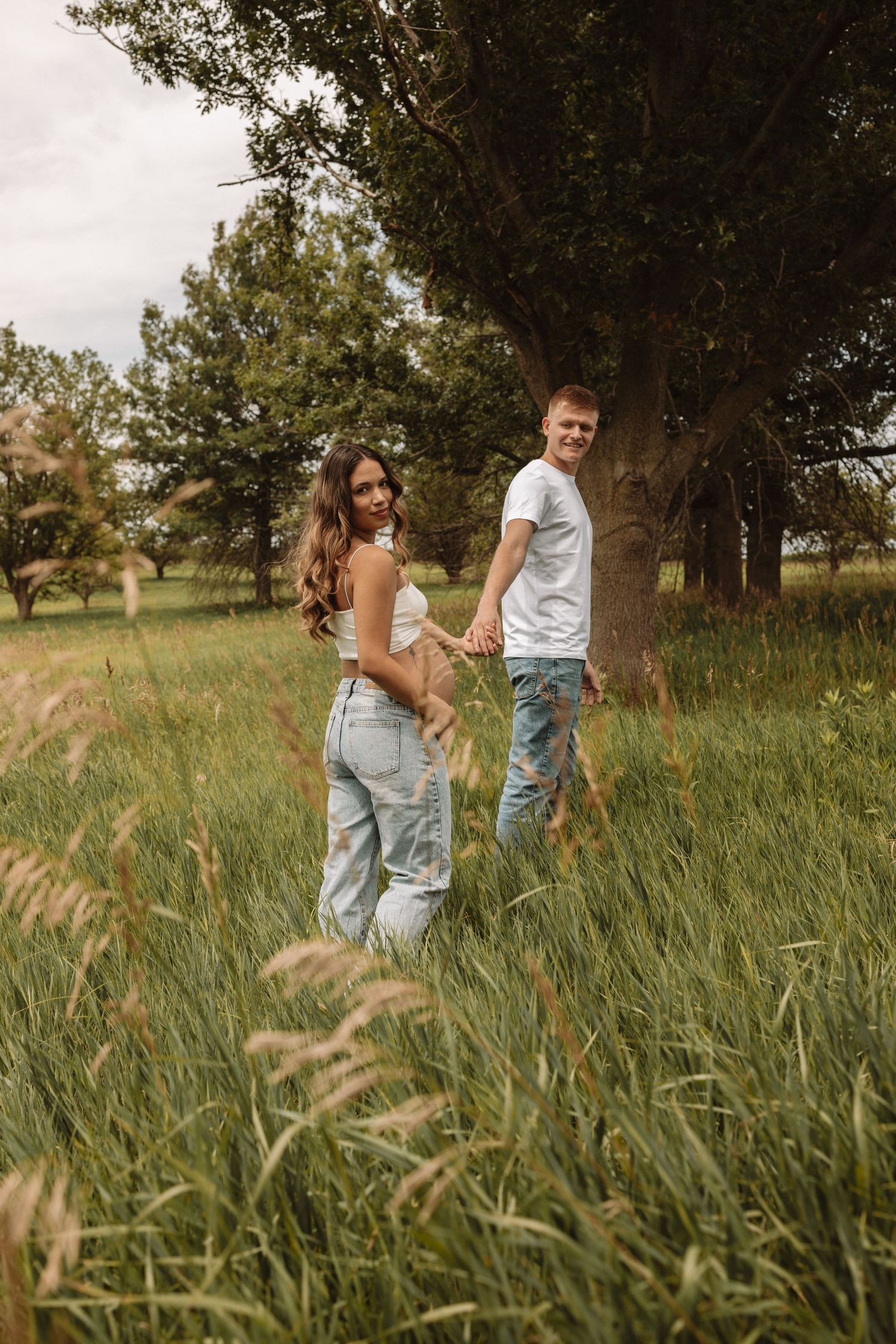 A couple stands close together in a rustic meadow with tall grass and trees on a warm summer afternoon.