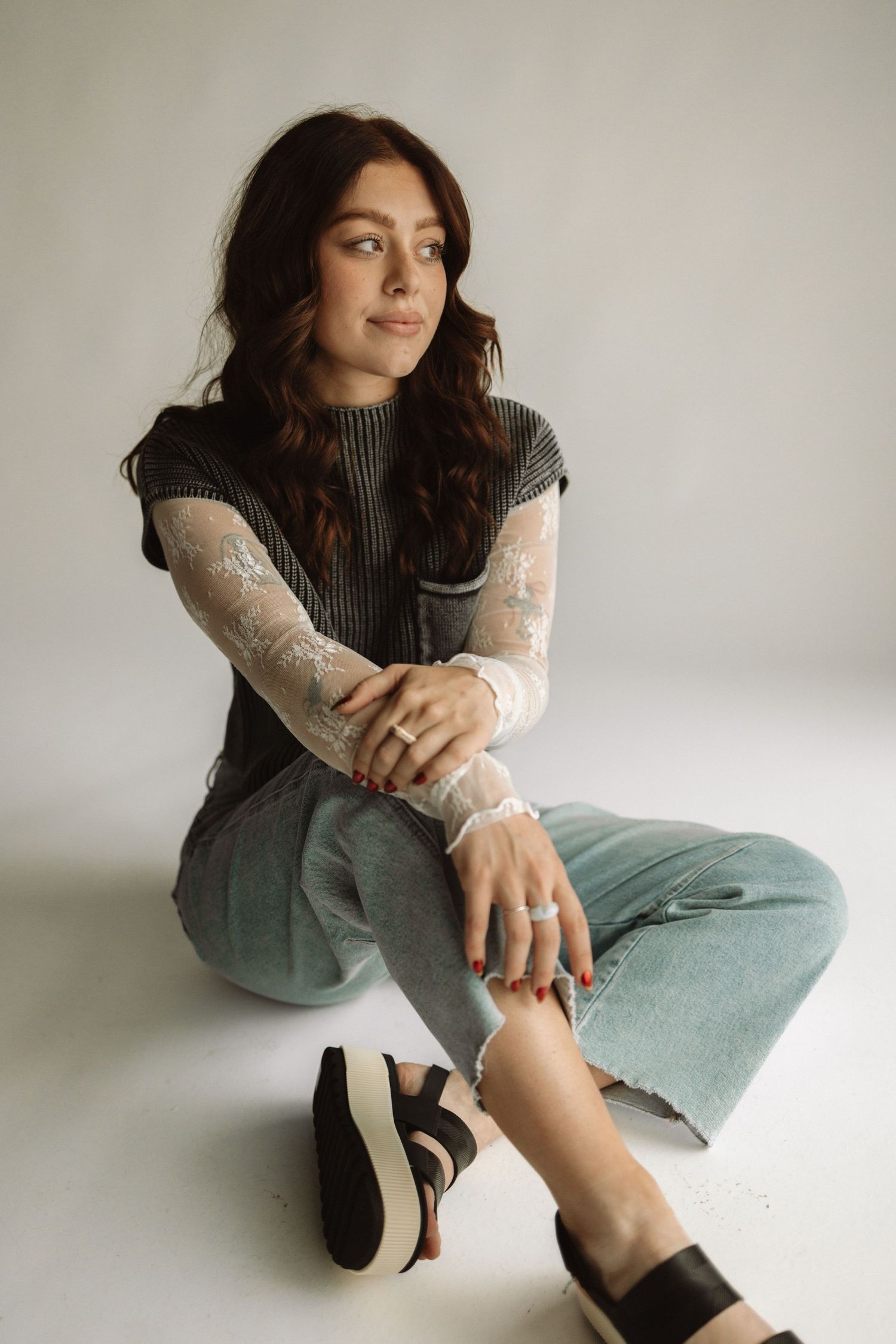 Person in gray sleeveless top and mint jeans sits casually on floor wearing black platform sandals in minimal setting.
