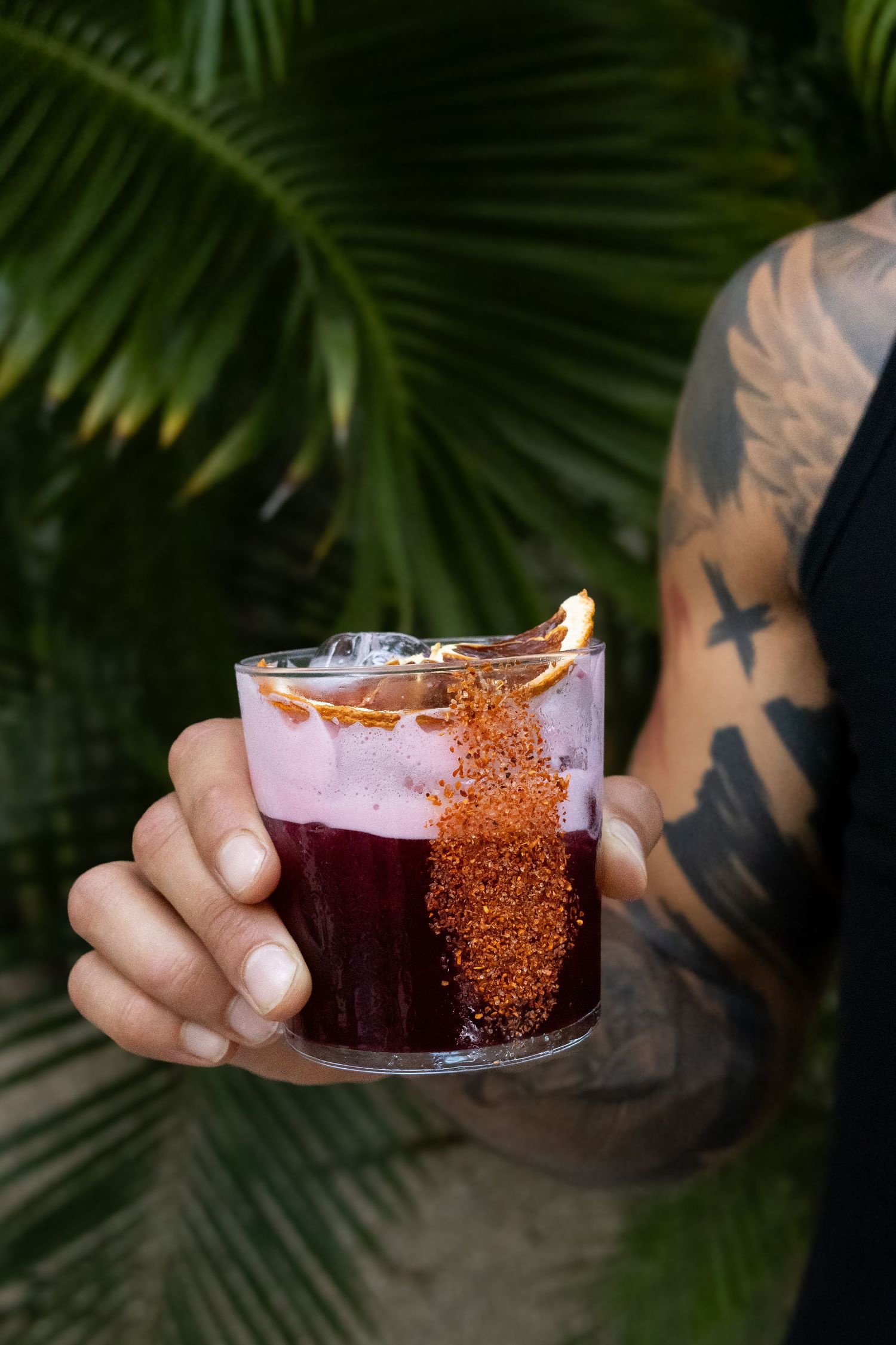 Fotografia gastronomica un cocktail de jamaica sostenido por una mano cerca del pecho de un hombre, por detras se ven palmeras
