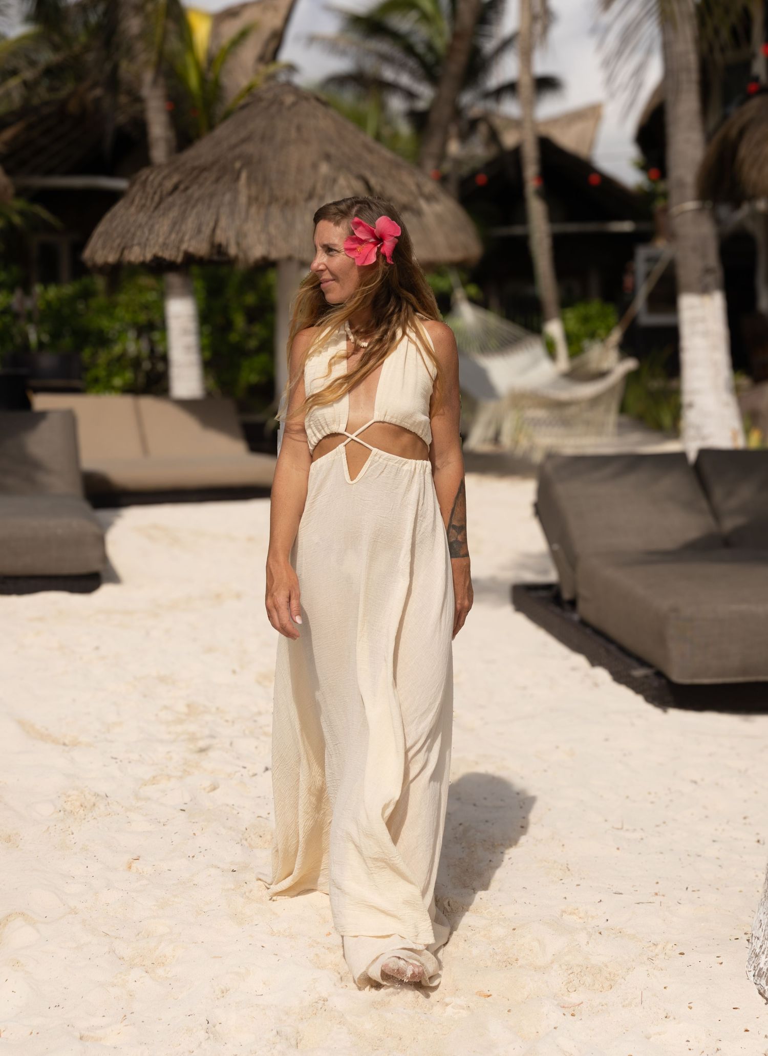 fotografia lifestyle. una modelo caminando en la playa con vestido blanco detras se ve parte del cheach club del hotel rustico