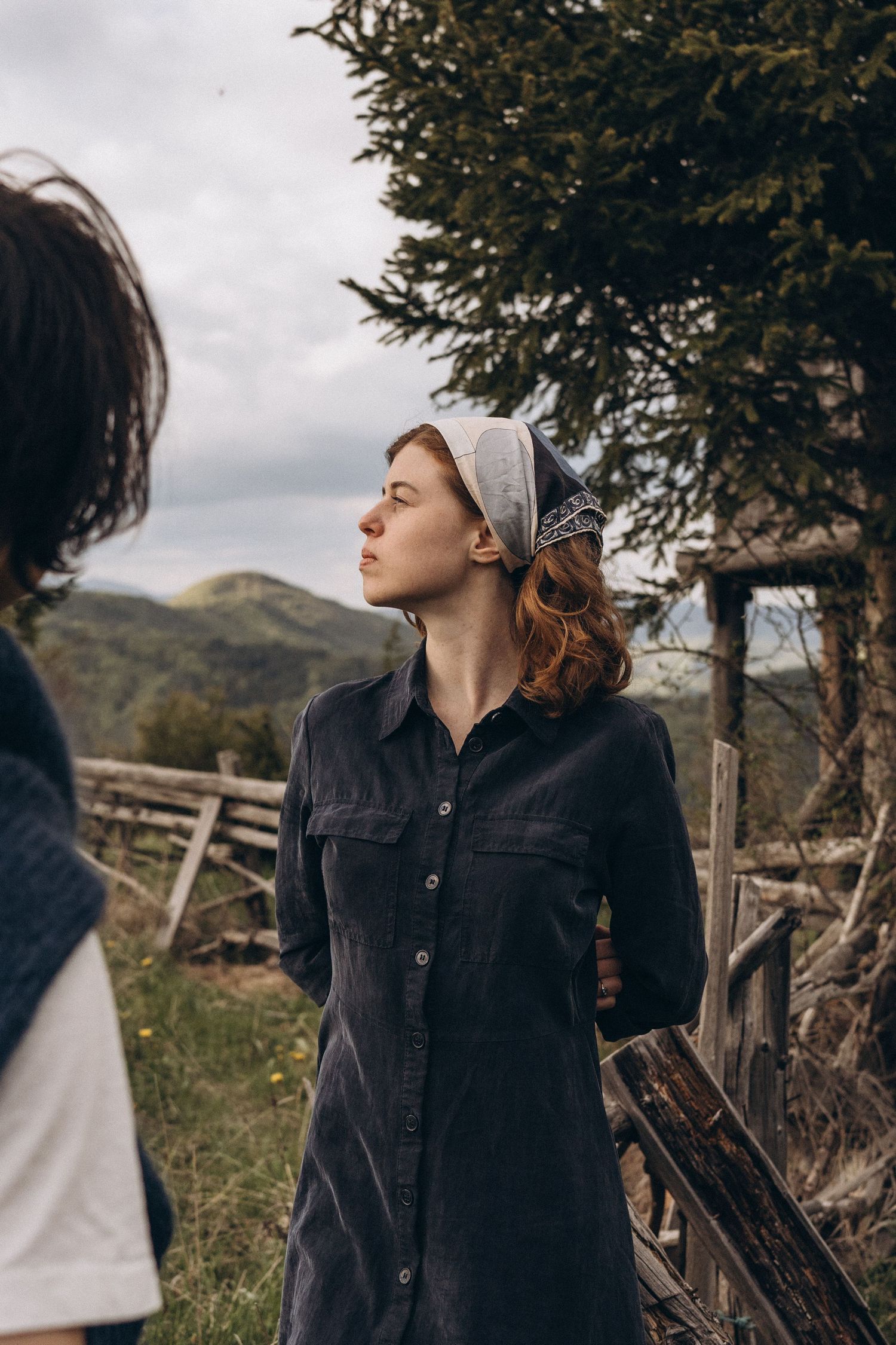 Person in dark jacket gazing upward while standing near wooden fence posts in nature.