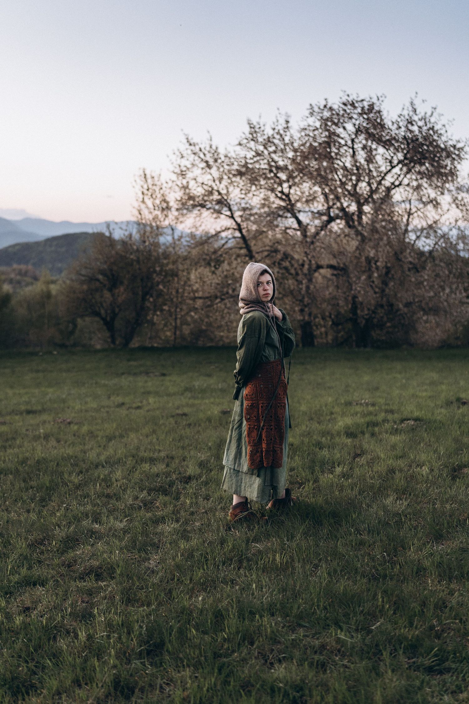 A person walks through a serene hillside meadow with bare trees against an overcast sky.