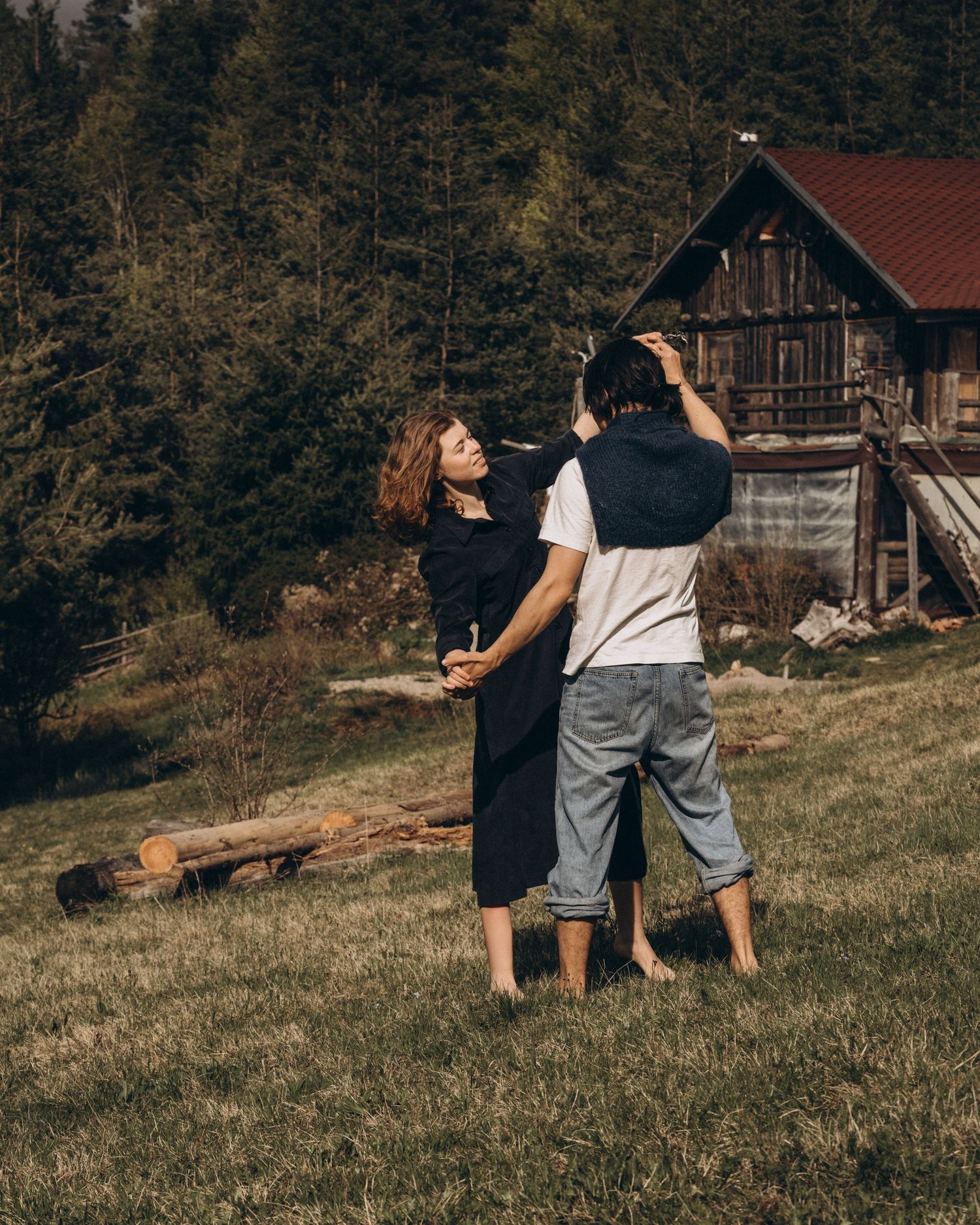 Two figures playfully interact on a grassy hillside with rustic cabins in the background.