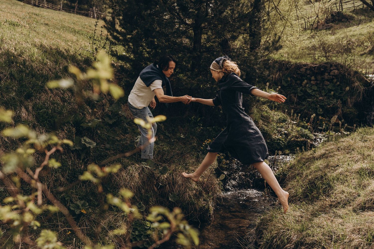 Two people helping each other cross a stream in a lush forest setting.