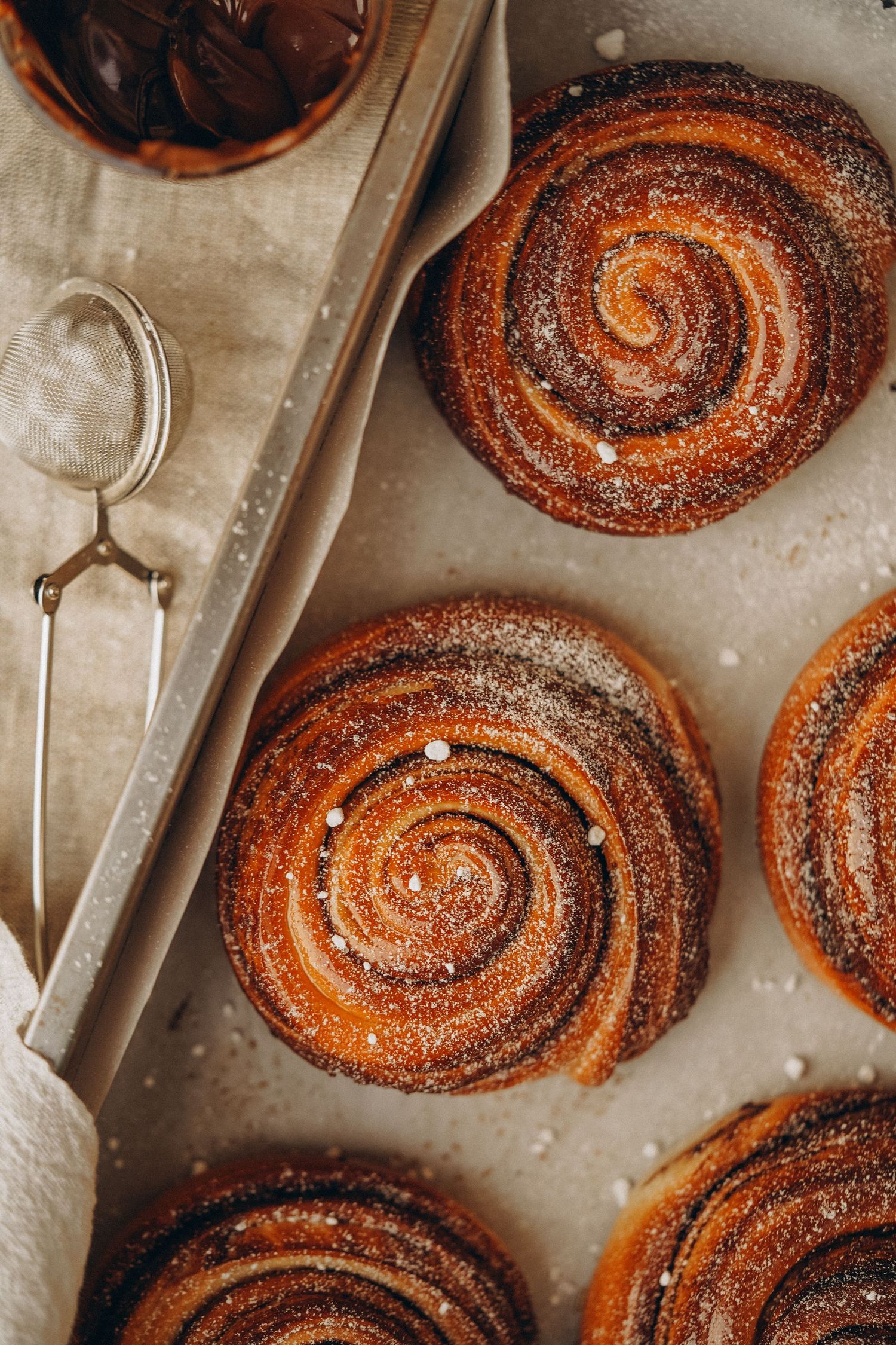 Freshly baked cinnamon rolls dusted with sugar on a baking sheet with serving utensils.