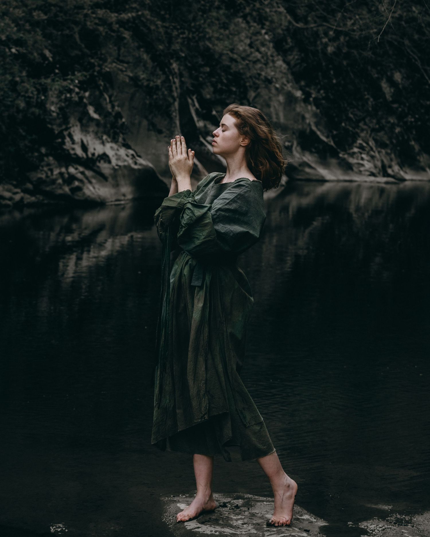 Dramatic dark photo of someone in black clothing posed by a lake.
