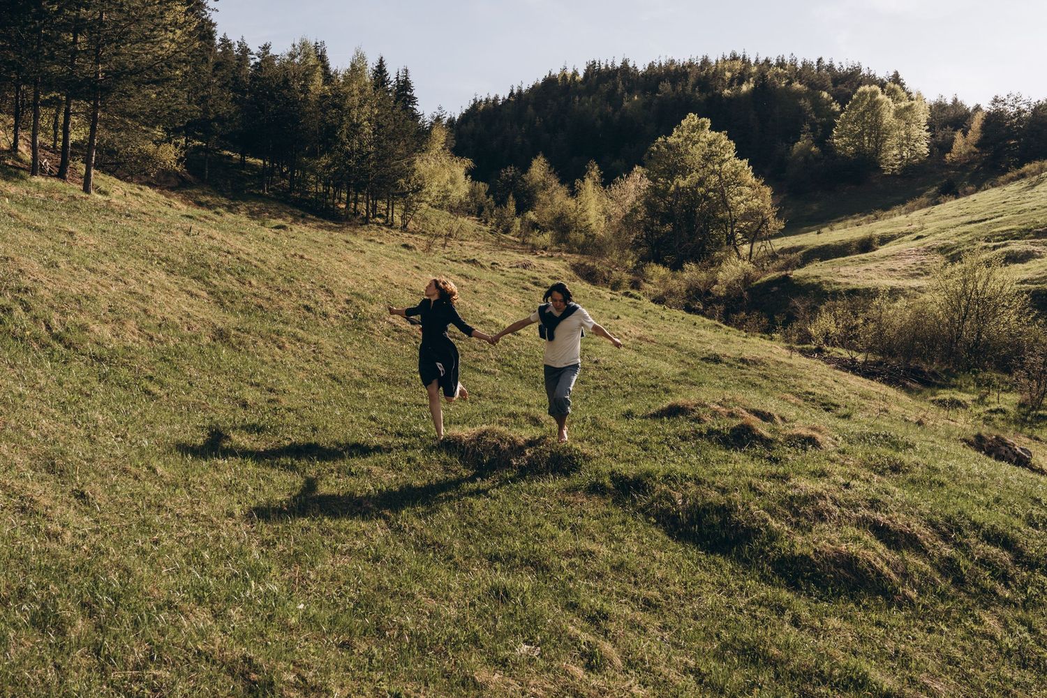 Two people running hand in hand down a grassy hillside surrounded by forest.