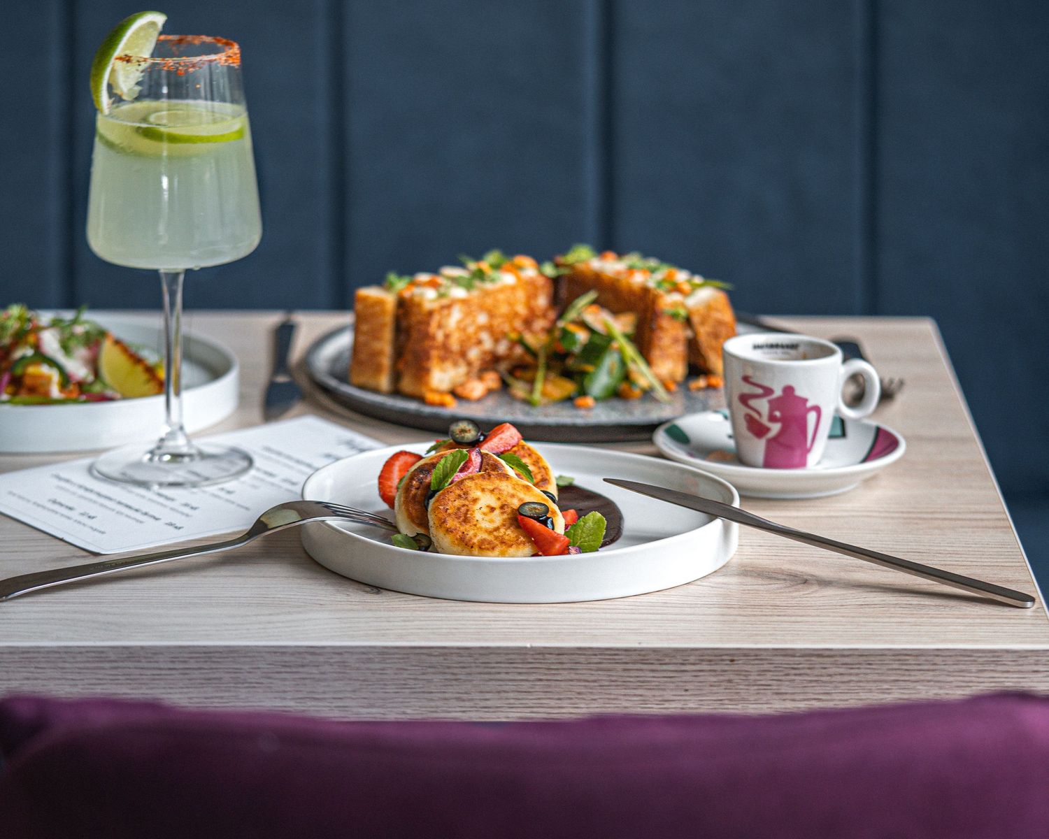 Restaurant table setting with cocktails and plated dishes against a dark blue wall.