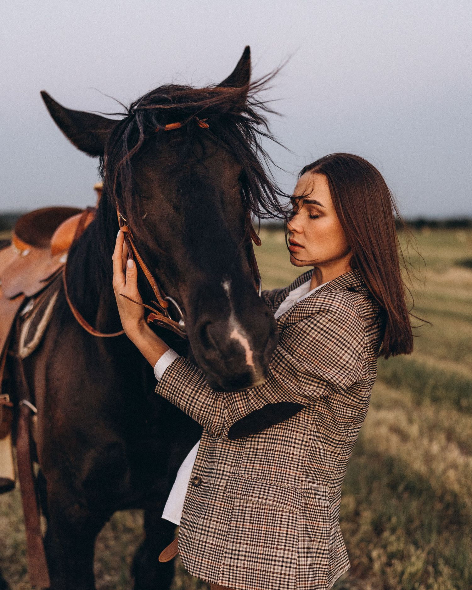 Person in plaid jacket tenderly interacts with dark horse in countryside setting.