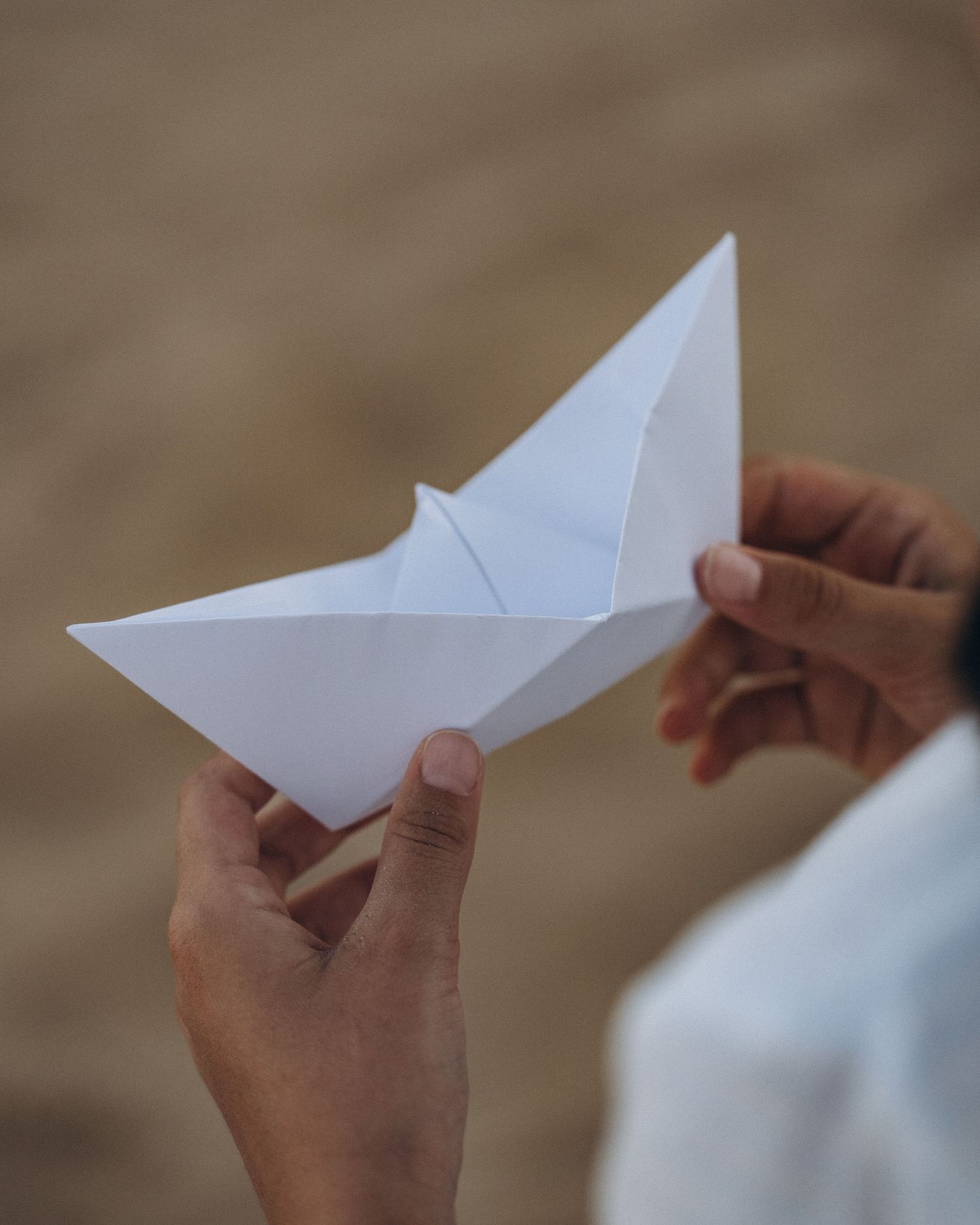 Hands holding and folding a white paper airplane against a soft brown background.
