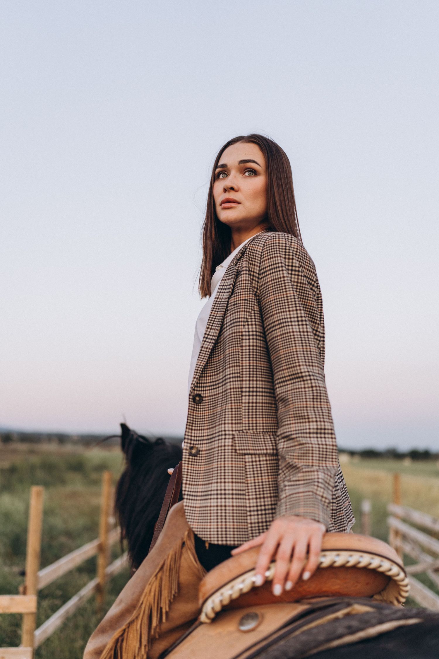 Person in plaid blazer sitting on horseback against rural landscape.