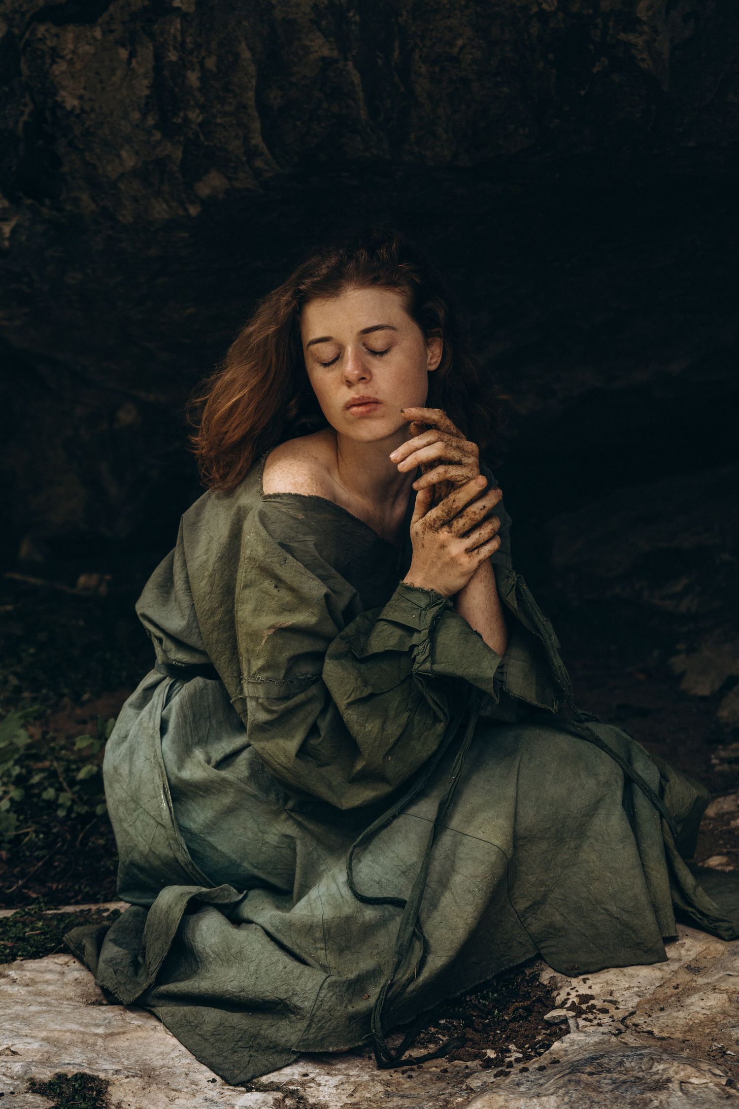 An artistic portrait showing someone in an olive dress hugging themselves against a dark cave wall.