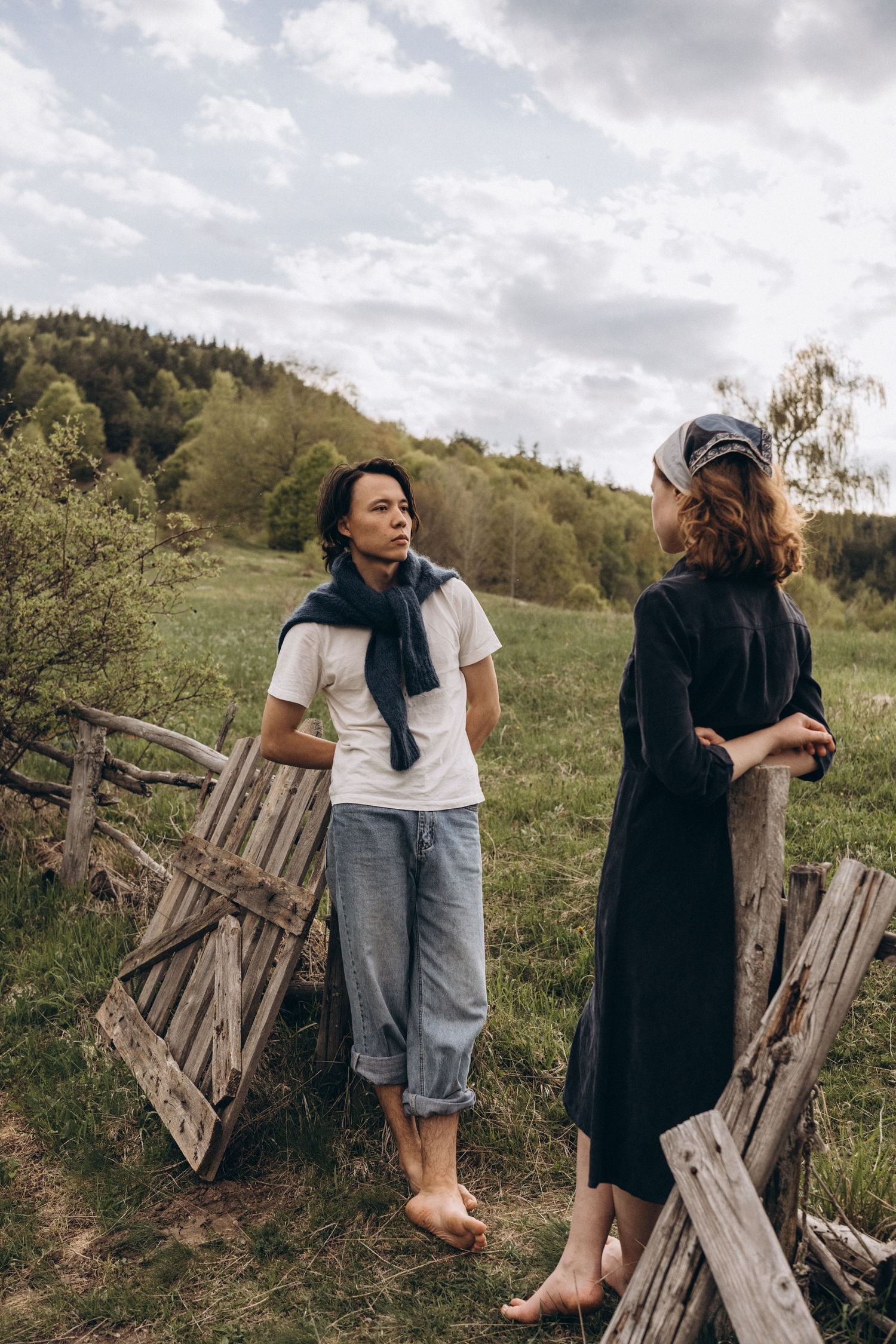 Two people in casual clothing interacting near rustic wooden fencing on a grassy hillside.