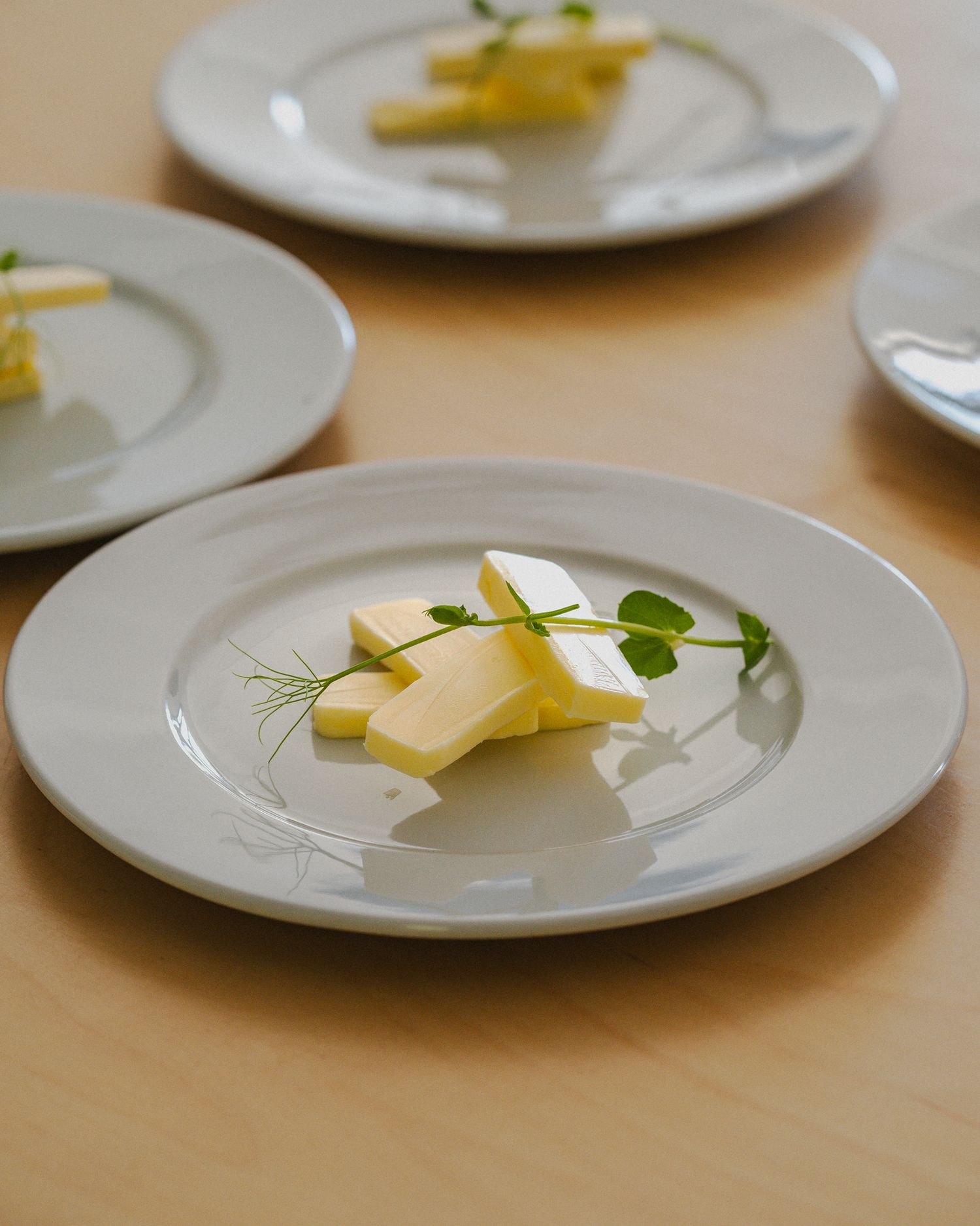 A row of white plates with thin slices of butter and fresh herbs arranged in a minimalist presentation.
