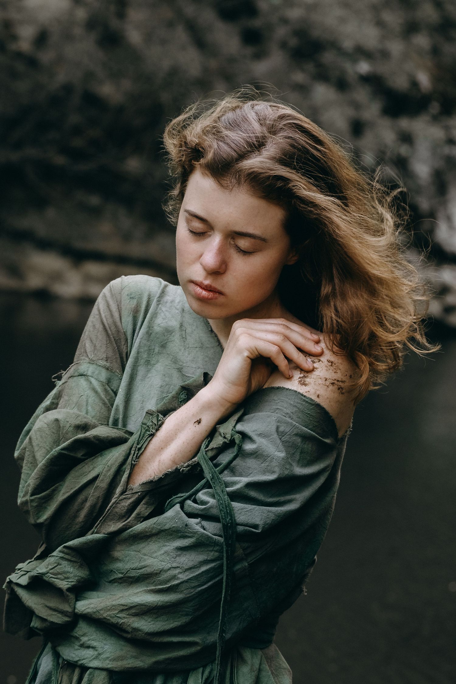 A moody portrait of someone in an olive green dress with windswept hair against a dark rocky background.