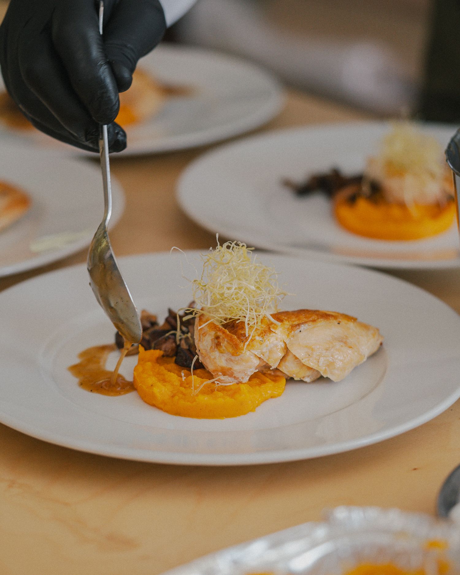 Chef in black gloves plates an elegant salmon dish with microgreens and orange puree on a white plate.