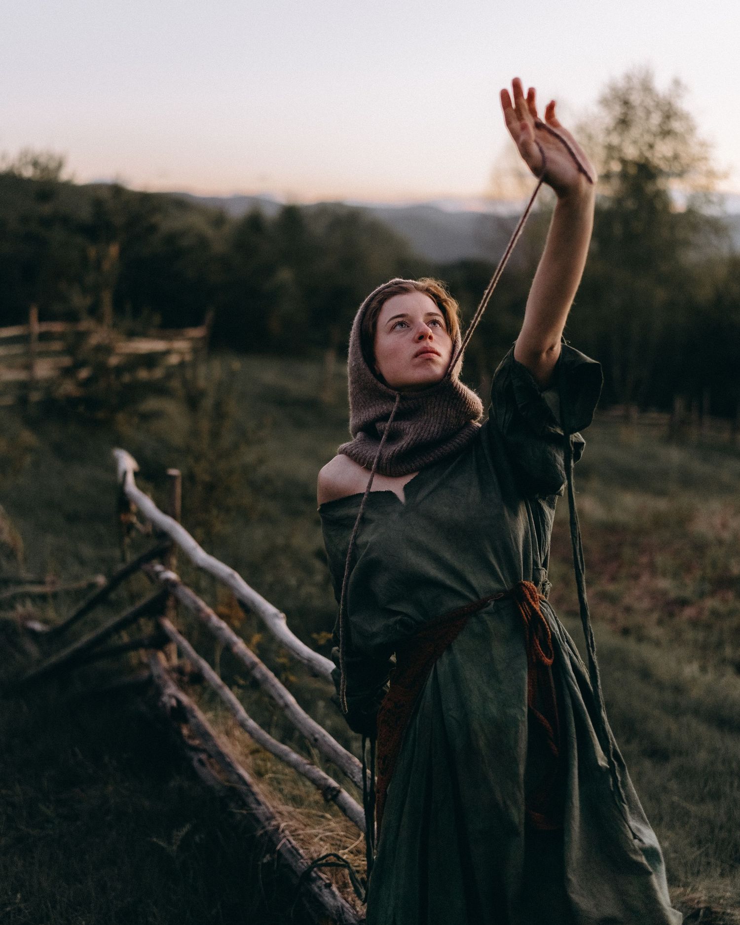 A figure in dark clothing reaches upward dramatically against a rustic outdoor landscape.