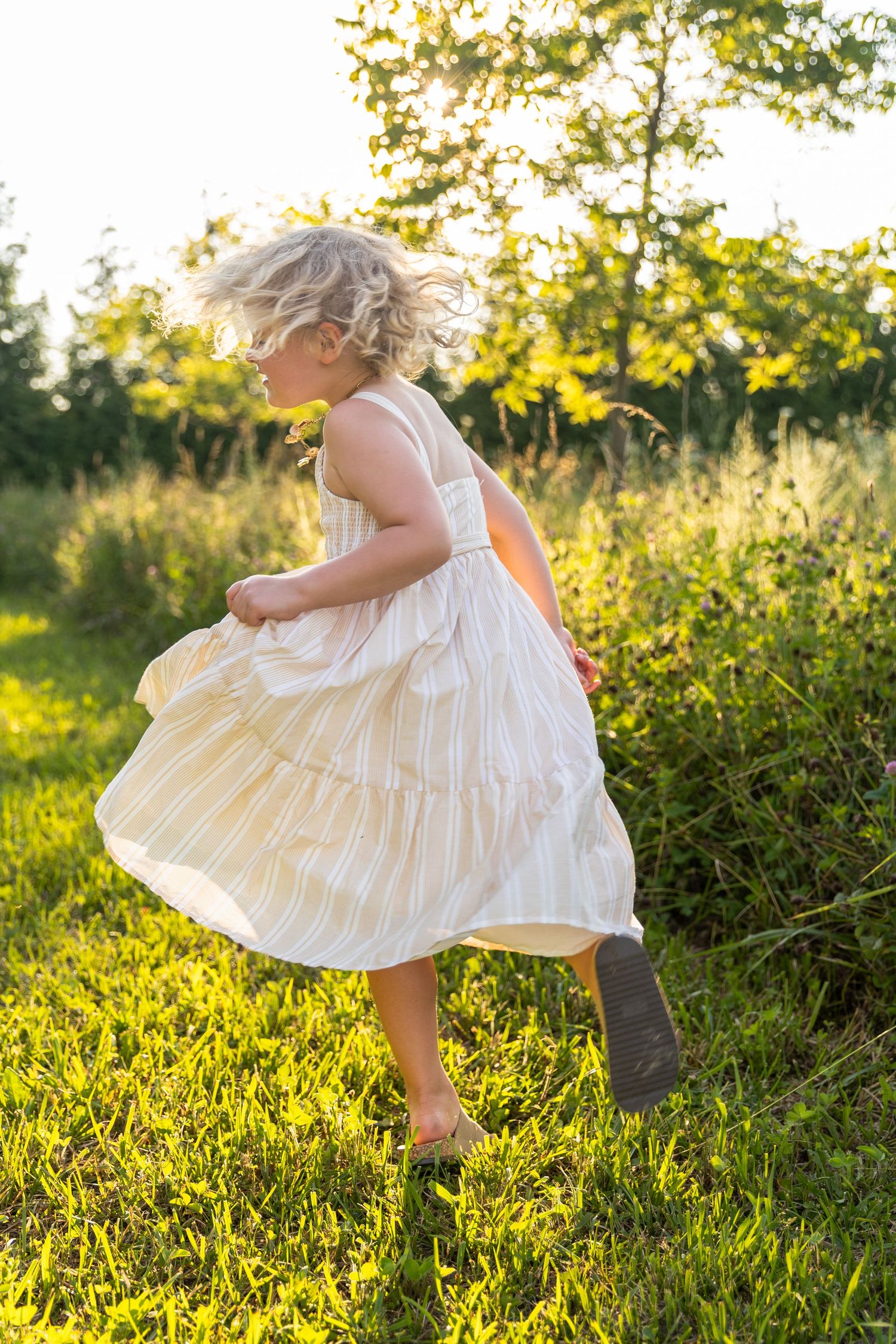 Young blonde girl in a flowing striped dress spinning barefoot in a sunlit meadow at golden hour.