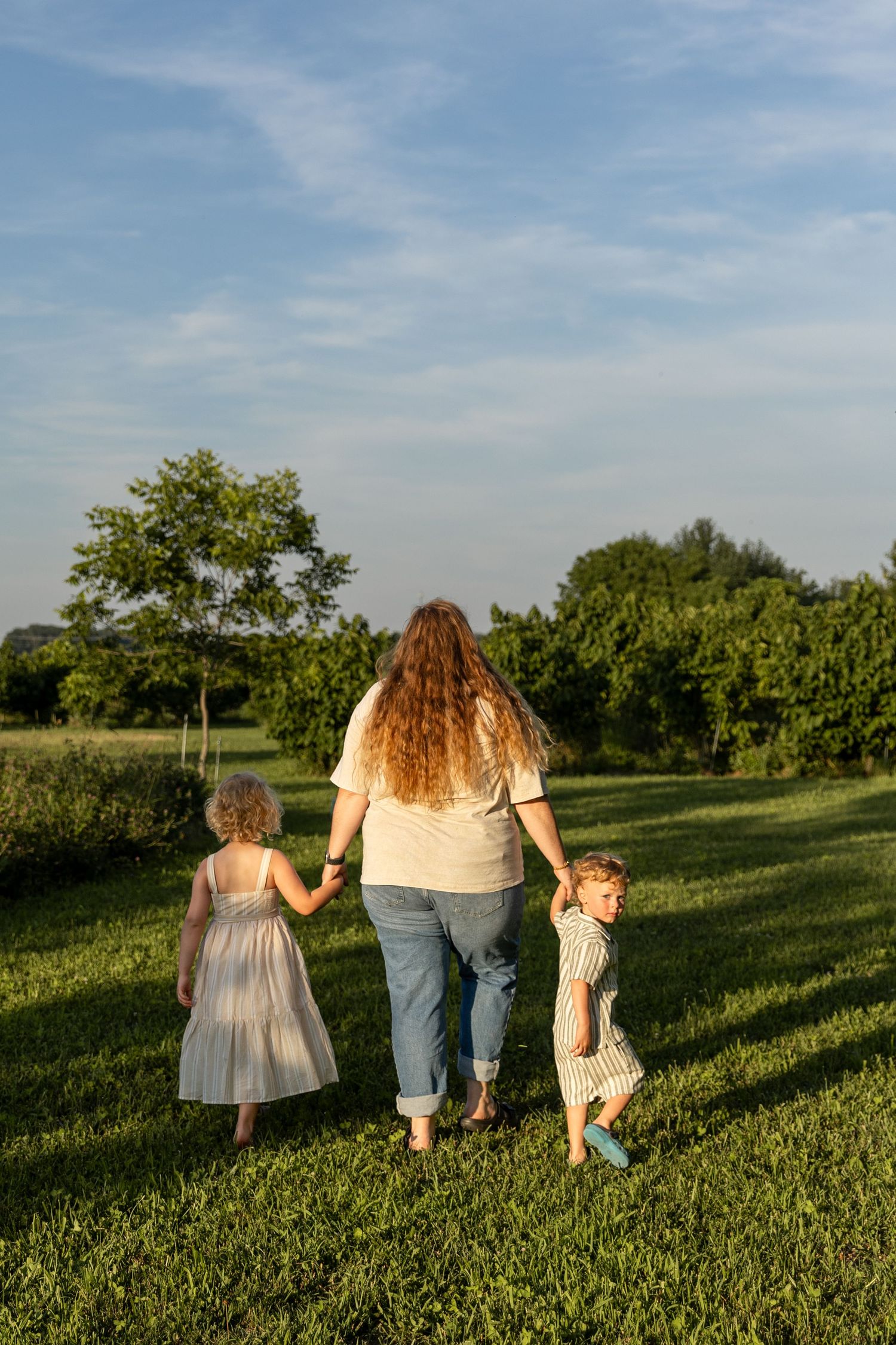 A mother walks hand-in-hand with her two young children across a lush green field at sunset.