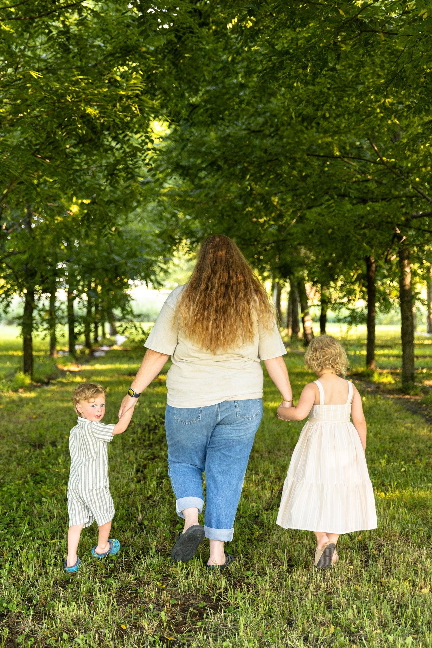 Mother walking hand-in-hand with two young children through green tree-lined path.