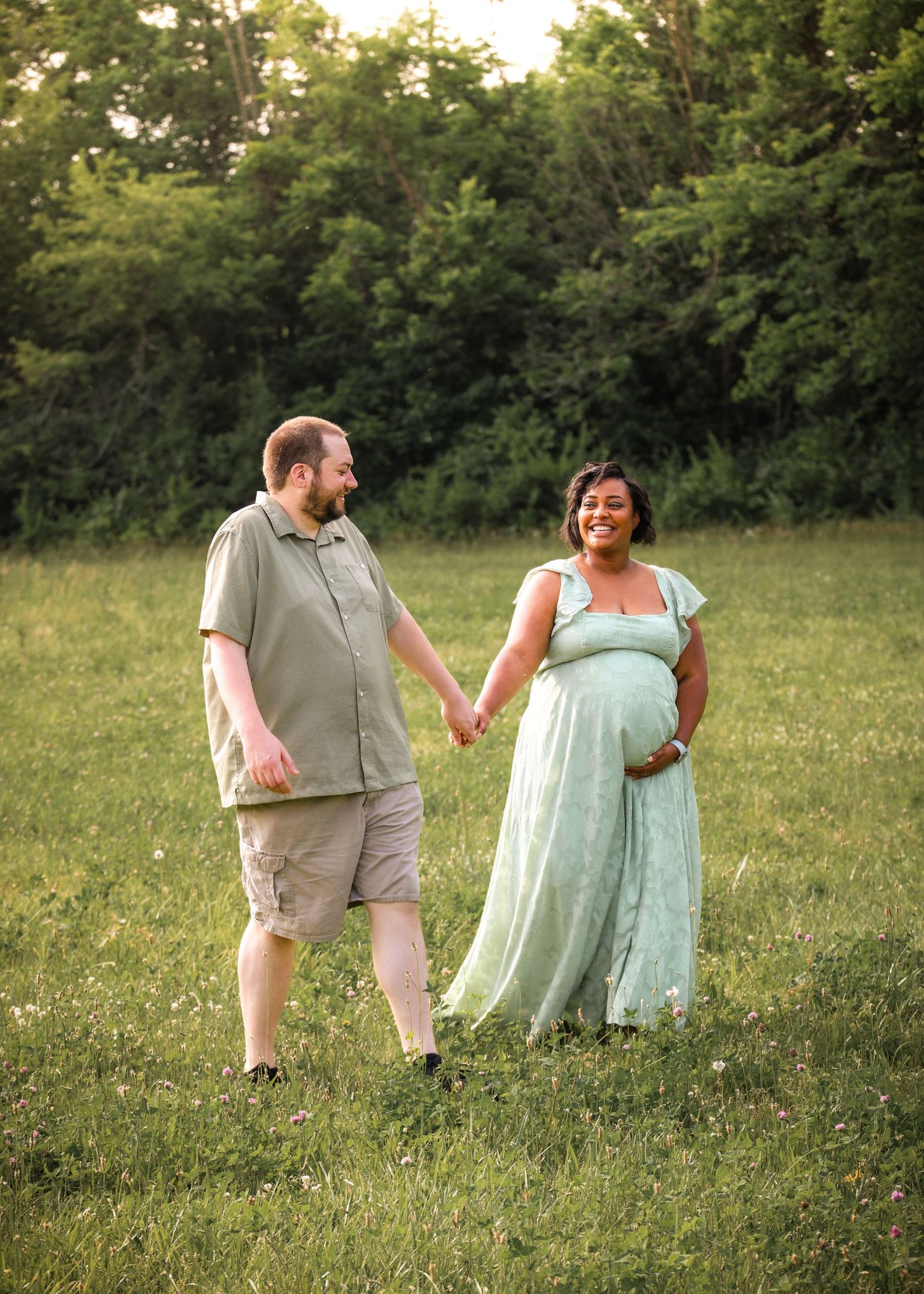 Happy pregnant couple holding hands and walking through a sunny green meadow.
