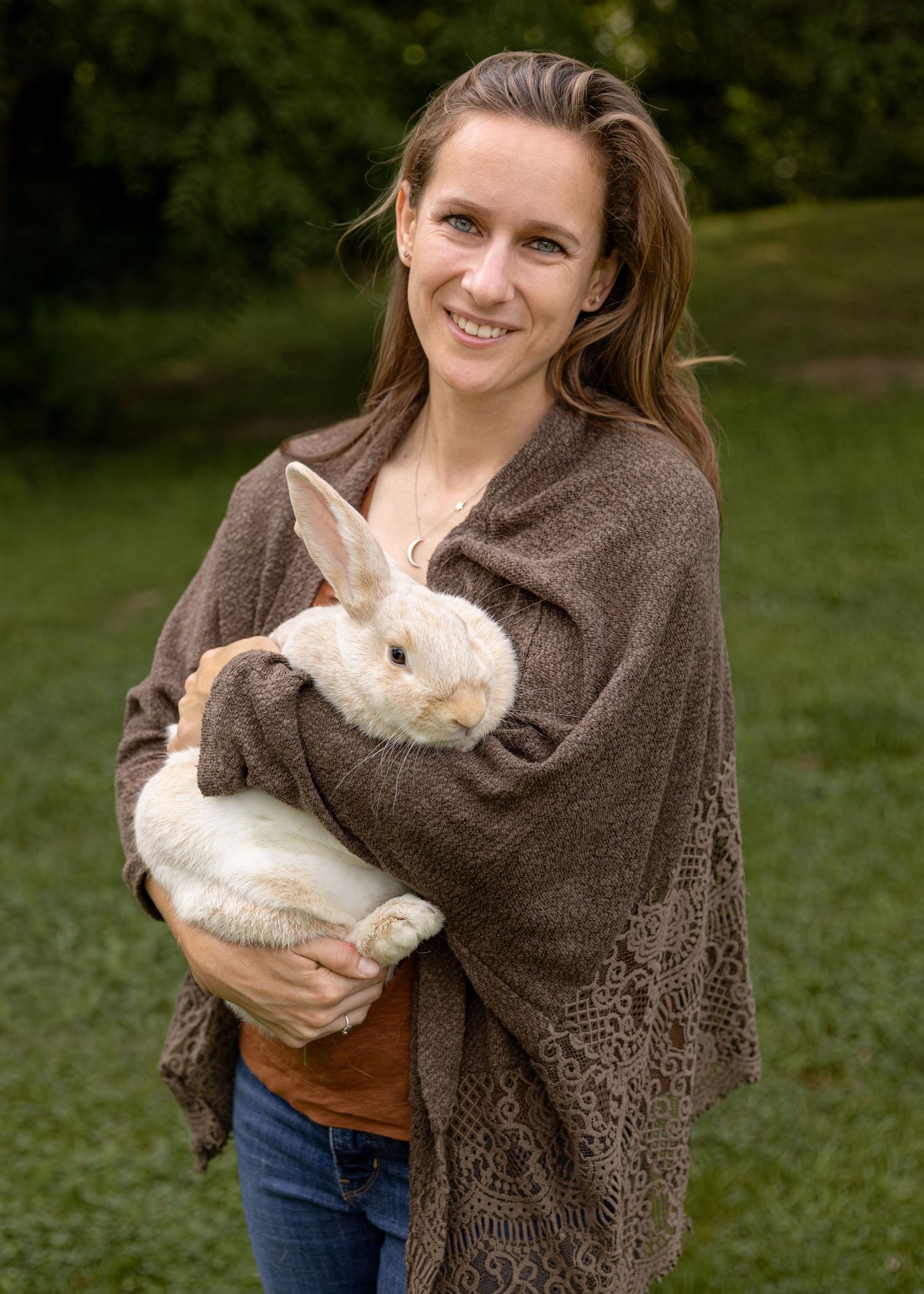 Smiling woman in brown shawl holding a Crème d’Argent rabbit outdoors on green grass.