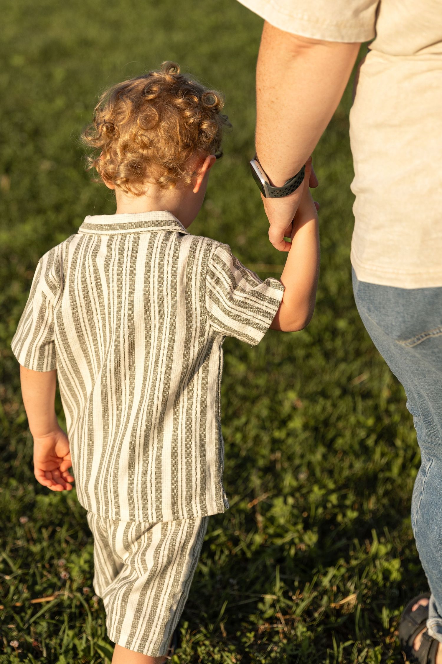 A young curly-haired toddler boy holds an adults hand while walking on green grass at sunset.