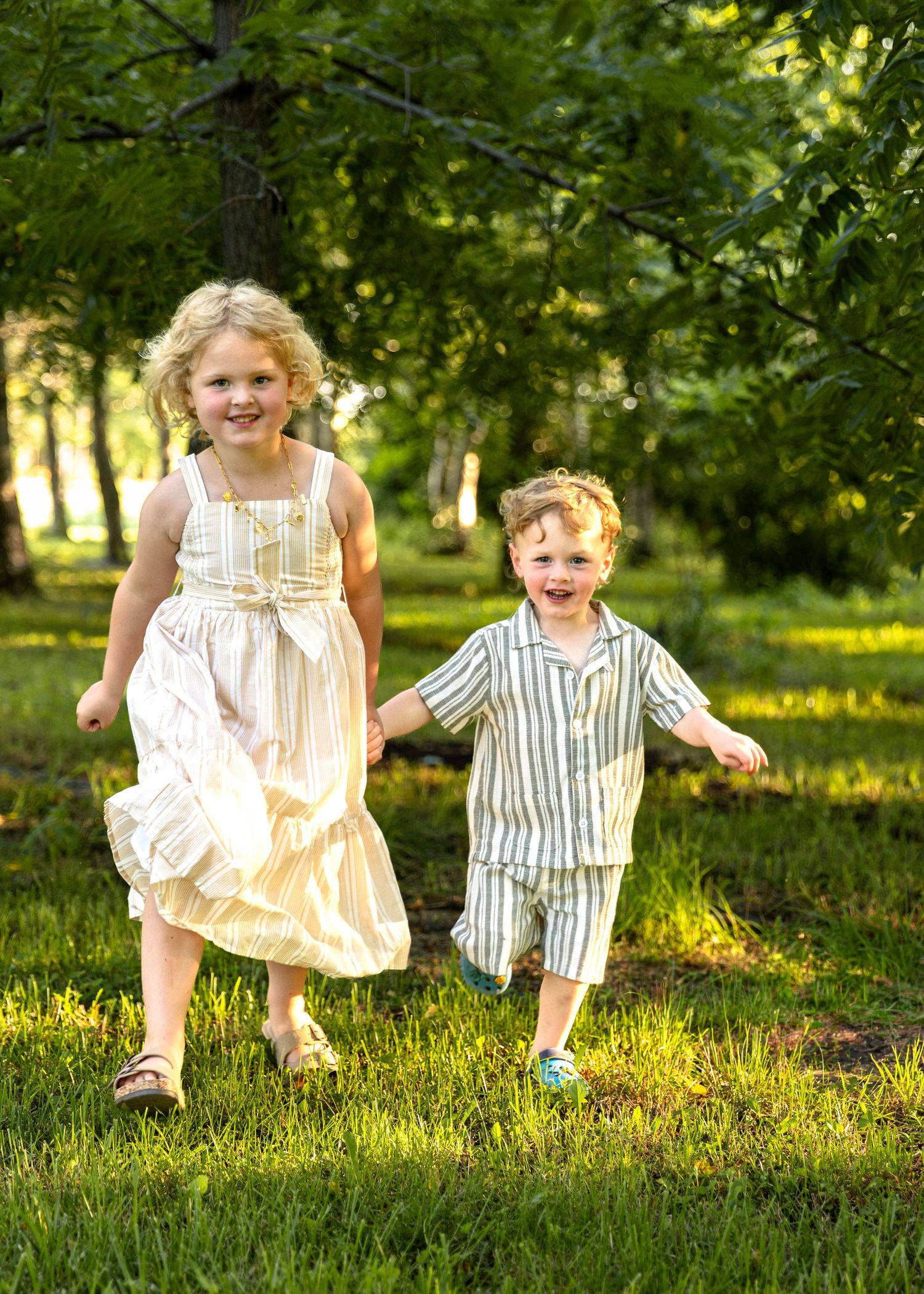 Two smiling young children in coordinating outfits run hand-in-hand through a bright green park.