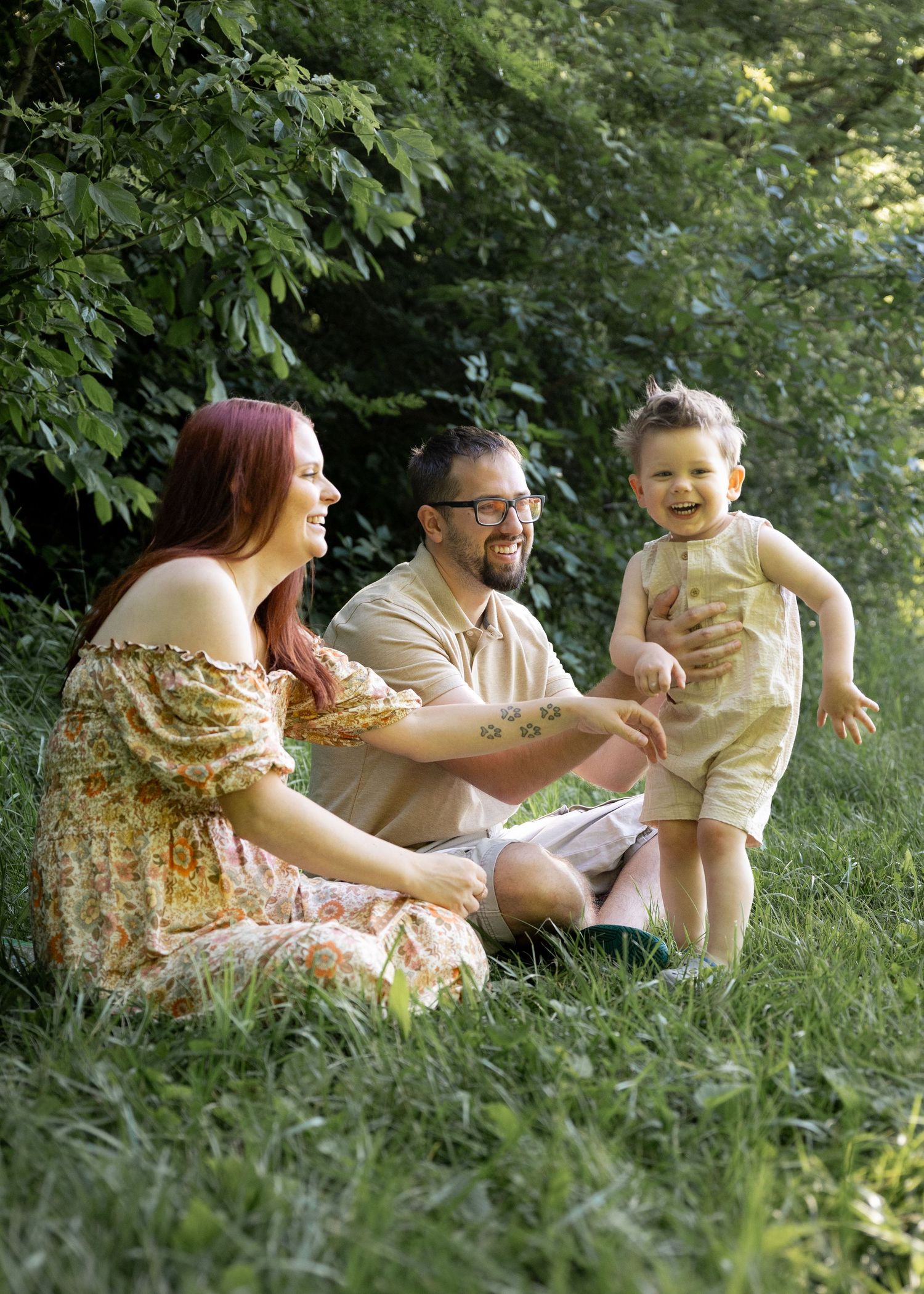 A joyful family of three sits on lush green grass, with a laughing toddler standing between smiling parents outdoors.