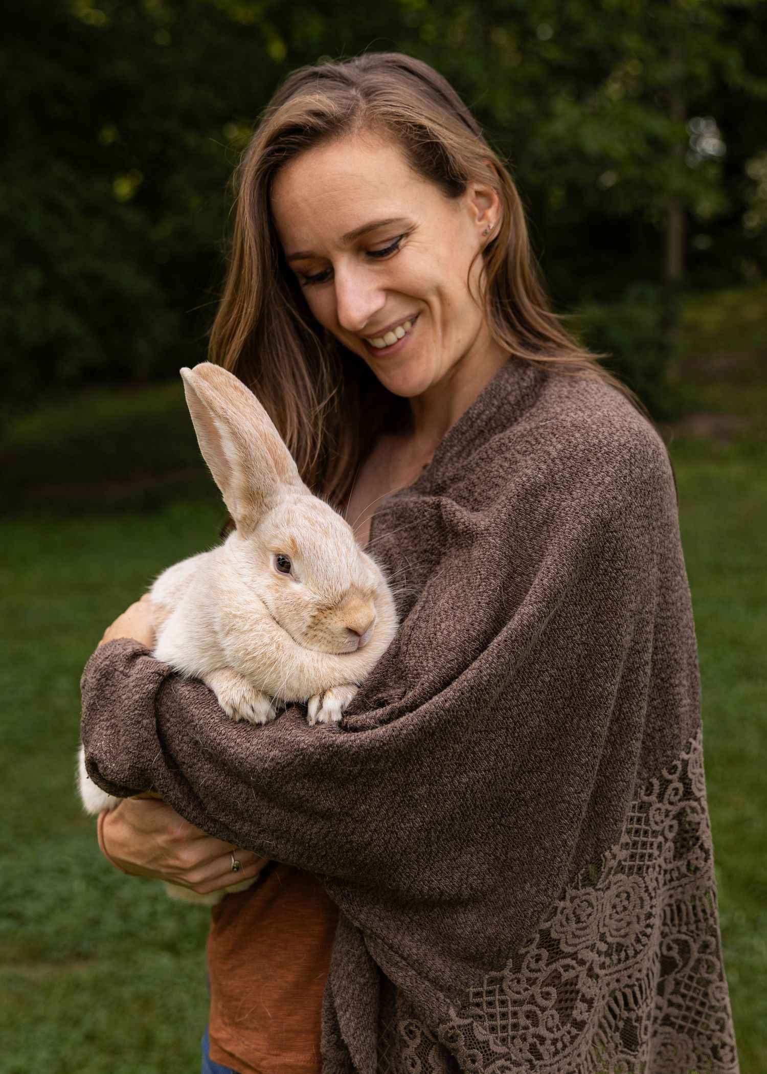 Smiling woman holding a Crème d’Argent rabbit wrapped in a brown shawl outdoors.