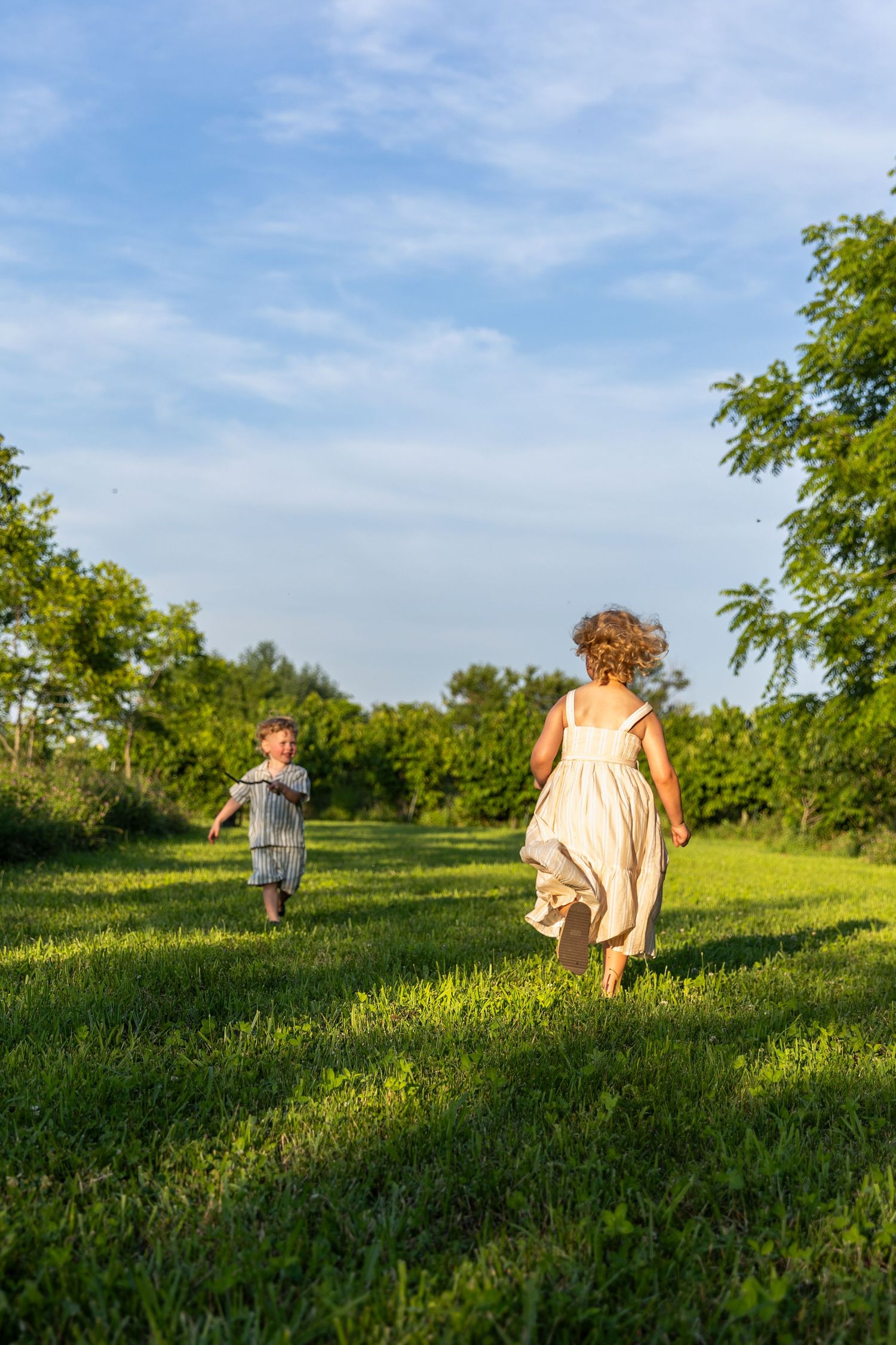 Two children running through lush green grass in a sunlit orchard on a warm summer day.