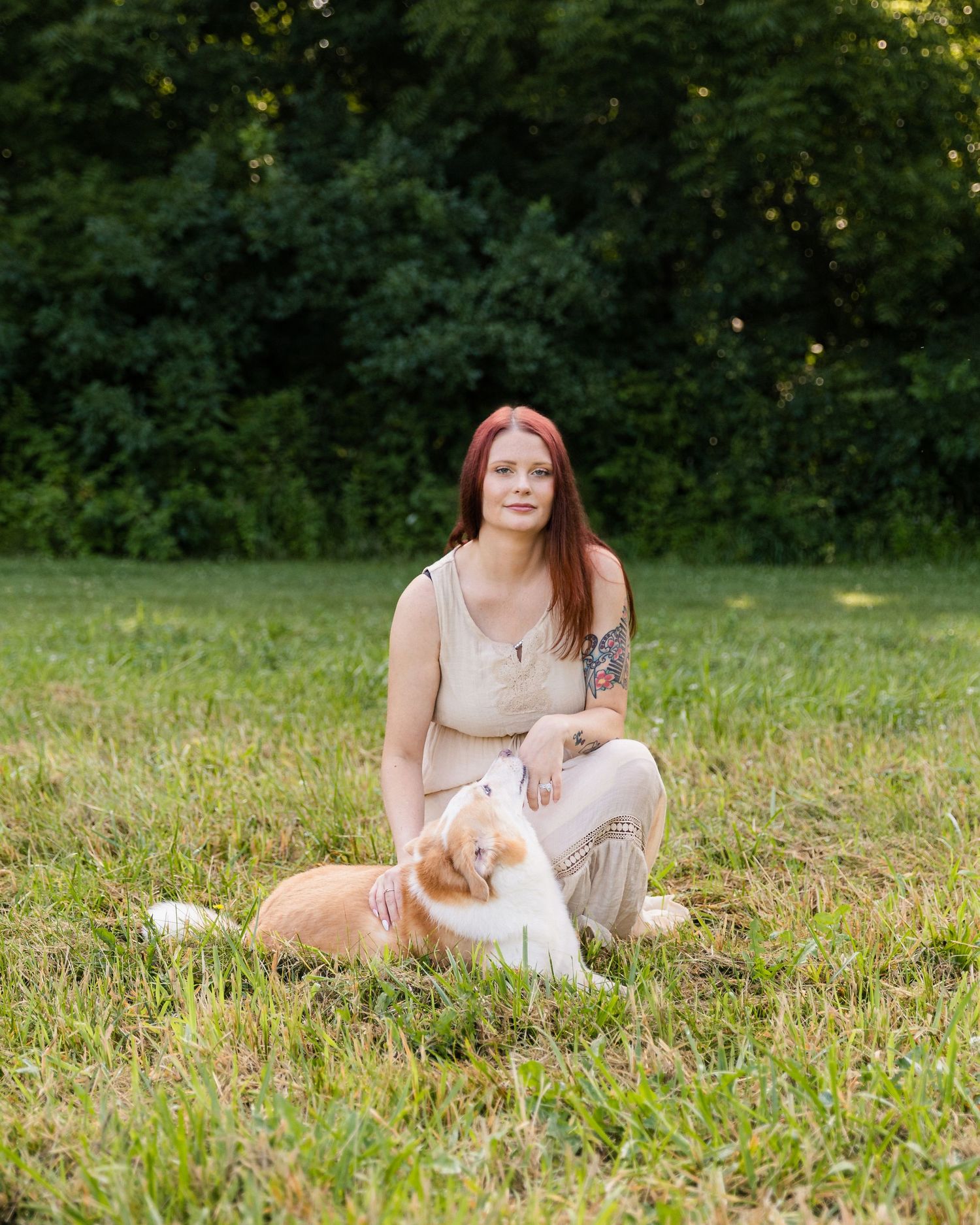 Red-haired woman sitting on grass with an orange and white dog in a park.