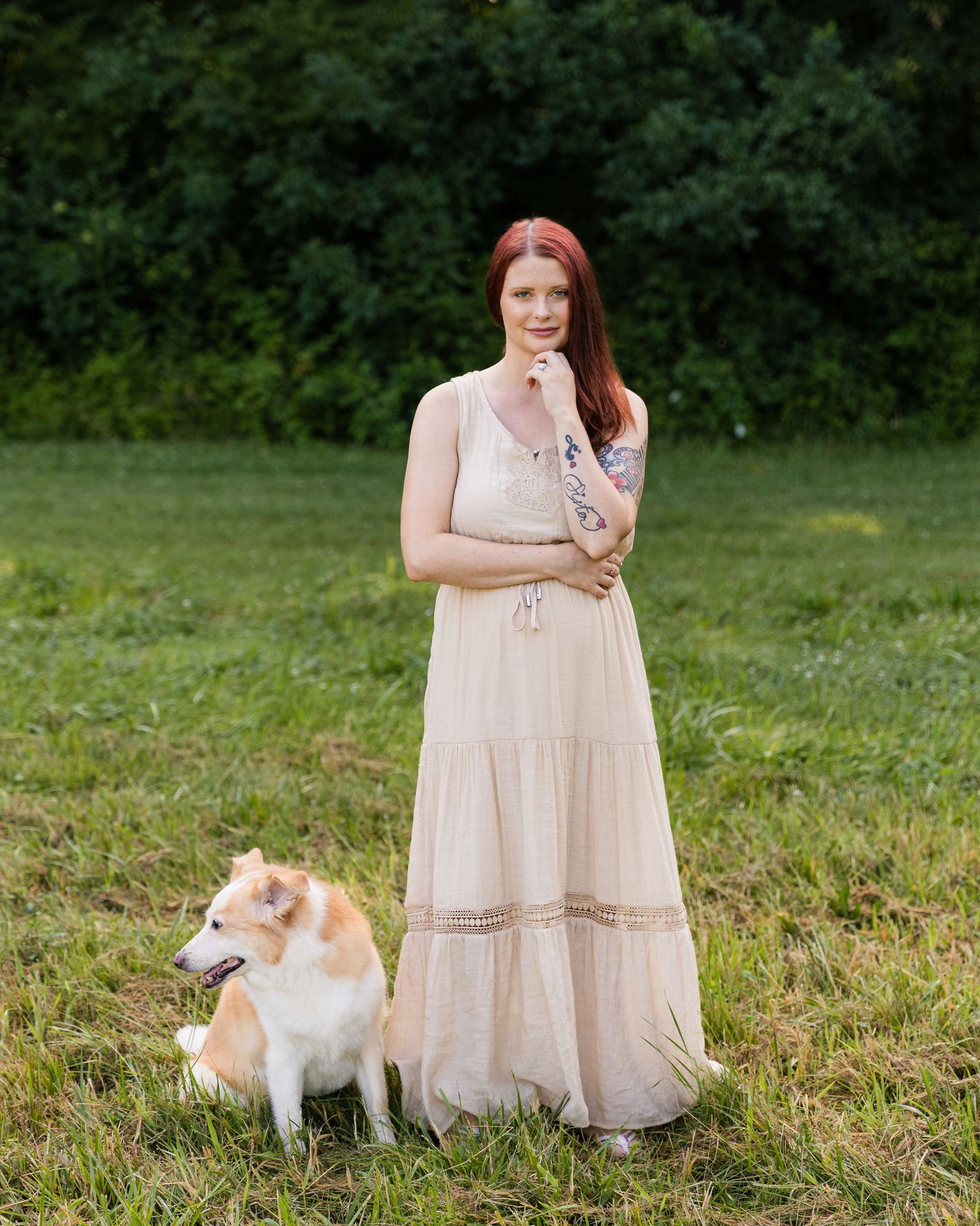 Red-haired woman in cream maxi dress standing outdoors with her dog on green grass.
