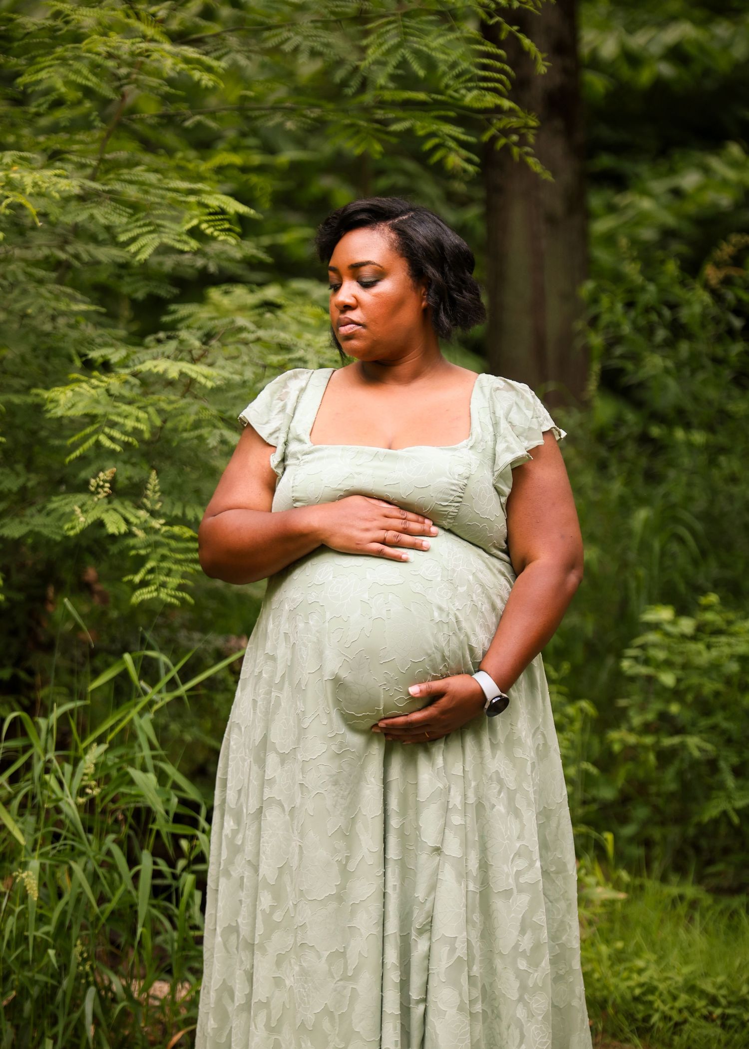 Pregnant woman in a sage green dress cradling her baby bump in a lush forest.