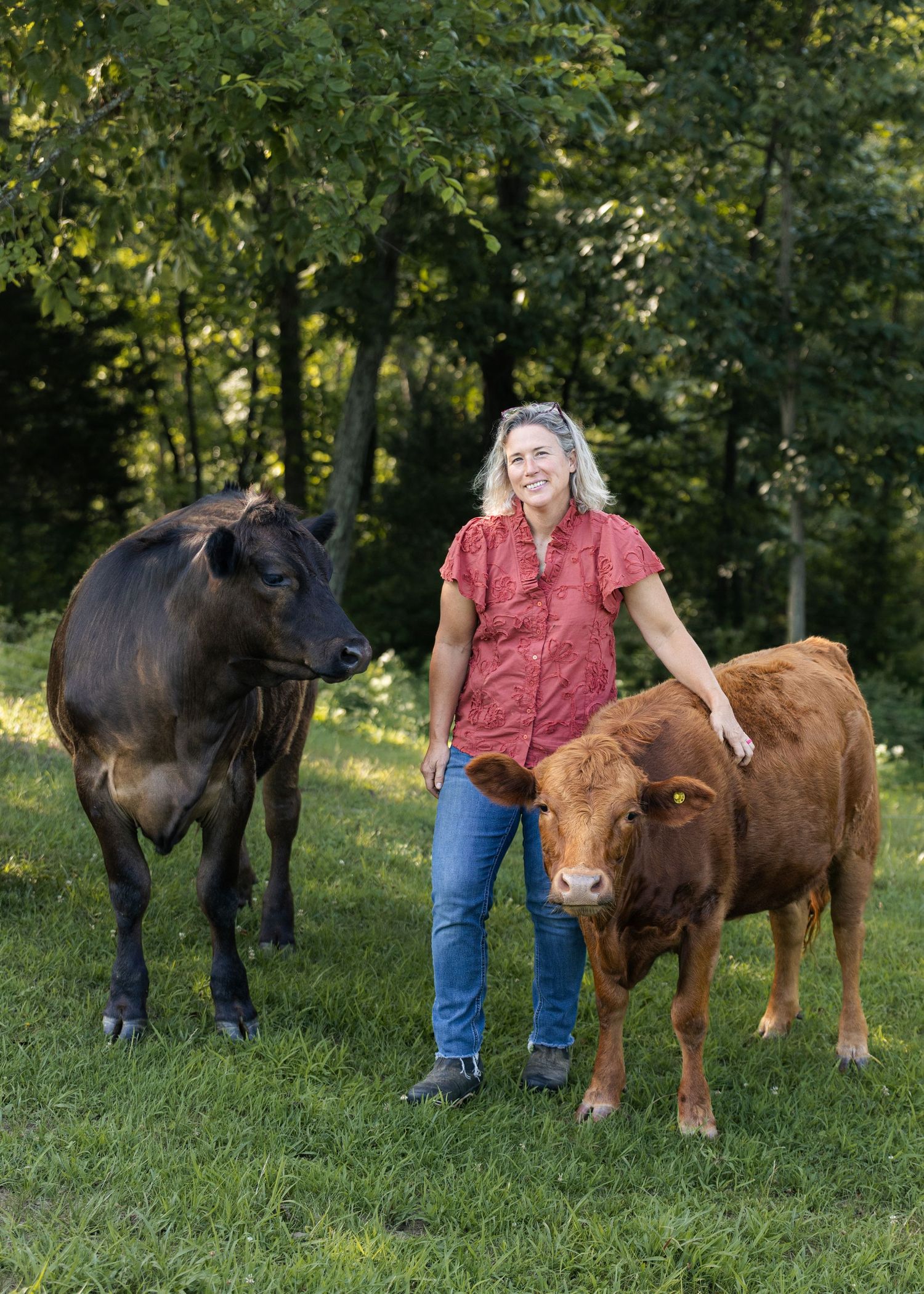 Woman smiling outdoors with two cows, a black and a red Dexter, on a lush green farm surrounded by trees.